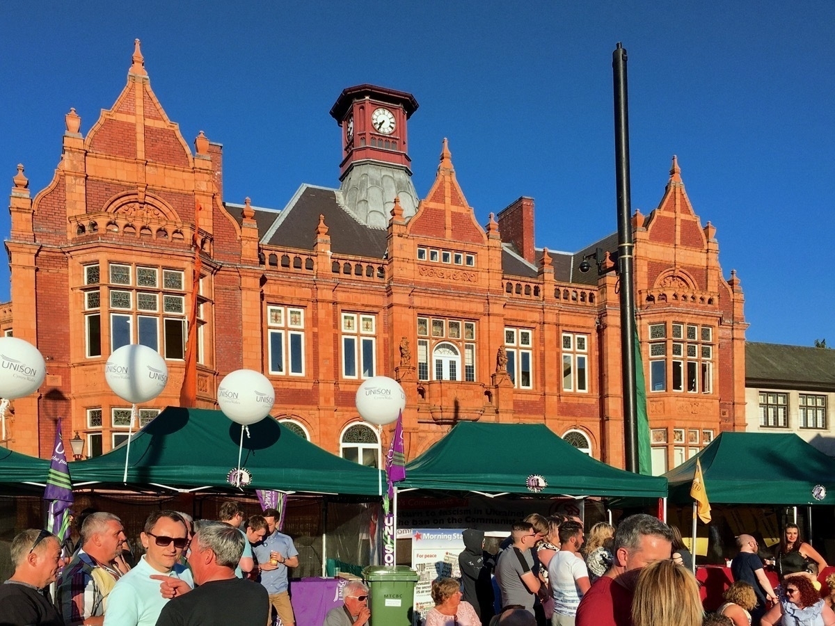 Large Victorian red brick building behind a row of market-type stalls. Groups of people in foreground, blue skies above.