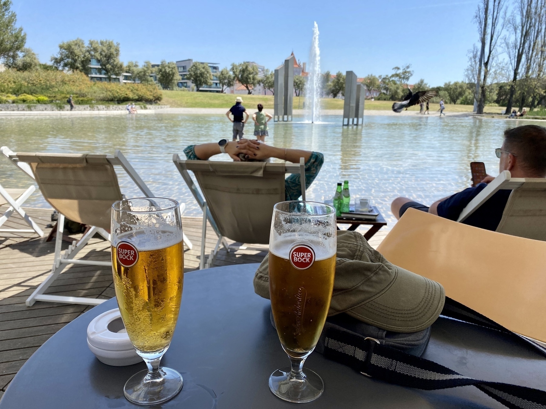 Two glasses of beer on a table with lake and fountain in background