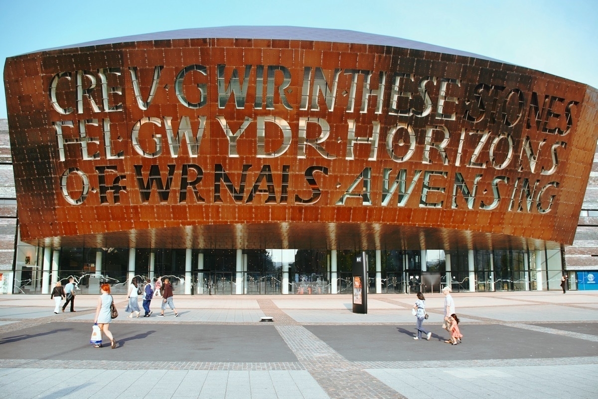 Curved copper plate frontage of a large theatre building with words formed by windows in the copper. Entrance doors below copper front, pedestrians strolling in the foreground