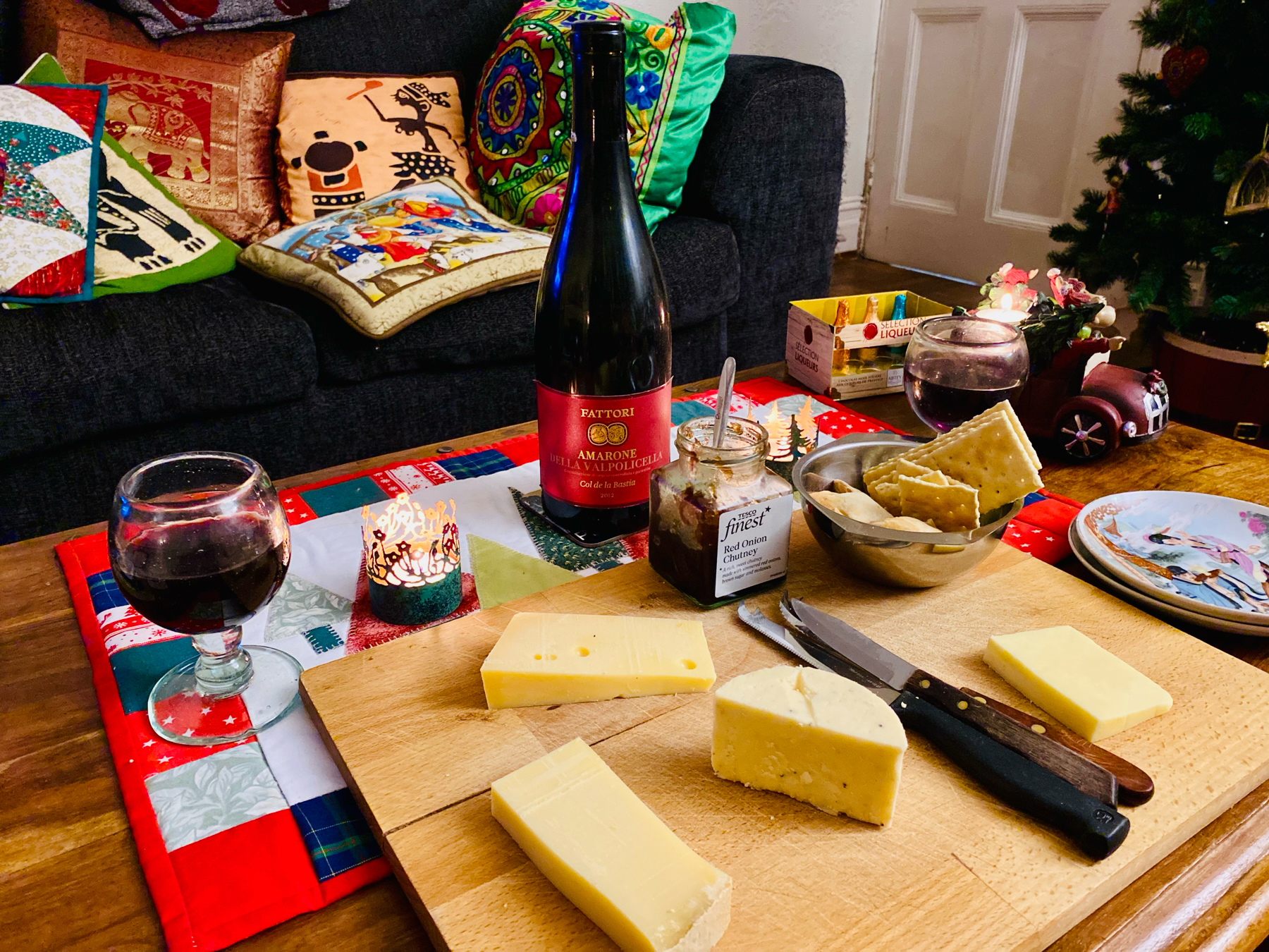 A cozy indoor scene with a festive atmosphere featuring a wine bottle, two glasses of red wine, assorted cheeses on a wooden board, crackers in a bowl, and a jar of red onion chutney. Decorative pillows, a Christmas tree.