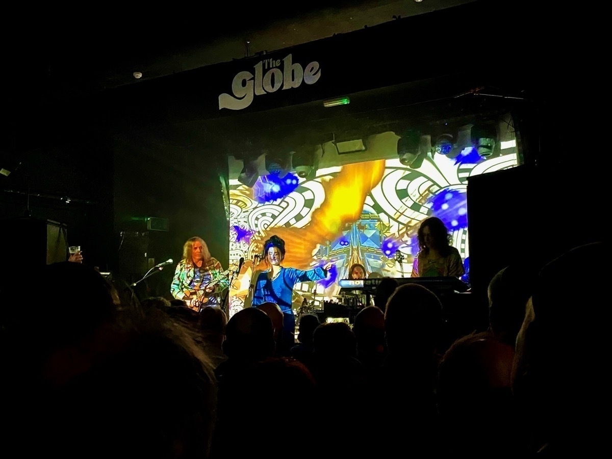 A live band performing on stage at The Globe, with colorful psychedelic projections in the background. Audience members are seen in the foreground, silhouetted against the brightly lit stage