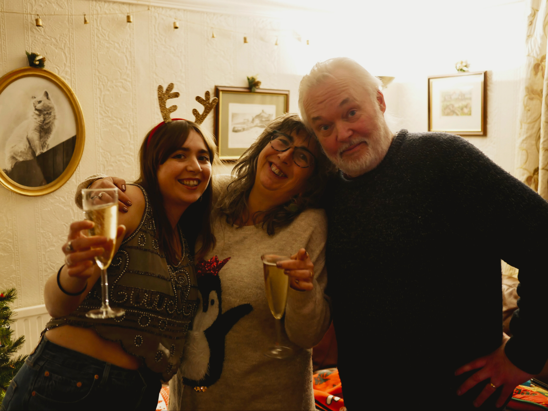 Three people smiling and posing for a photo indoors, likely during a festive gathering. The woman on the left is holding a glass of champagne, the woman in the middle is wearing glasses and wearing a holiday-themed jumper with a penguin.