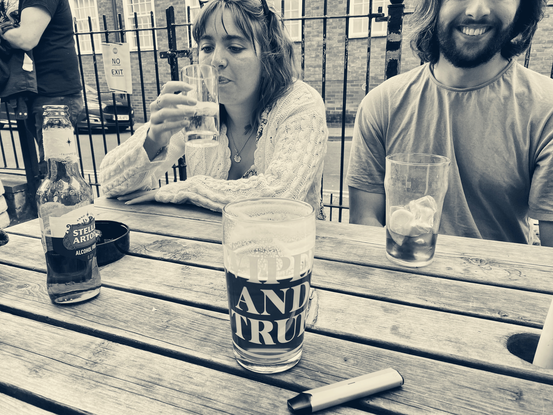 Monochrome picture of pint of beer on an outdoor pub table, woman drinking beer on opposite side of table