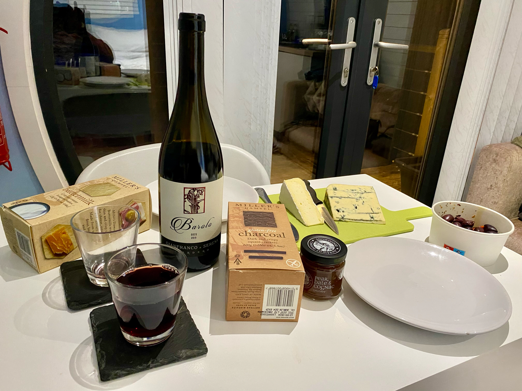 A table set with a bottle of Barolo wine, two glasses, a box of crackers, a plate of cheese, a bowl of olives, and a small jar of pear and date chutney.