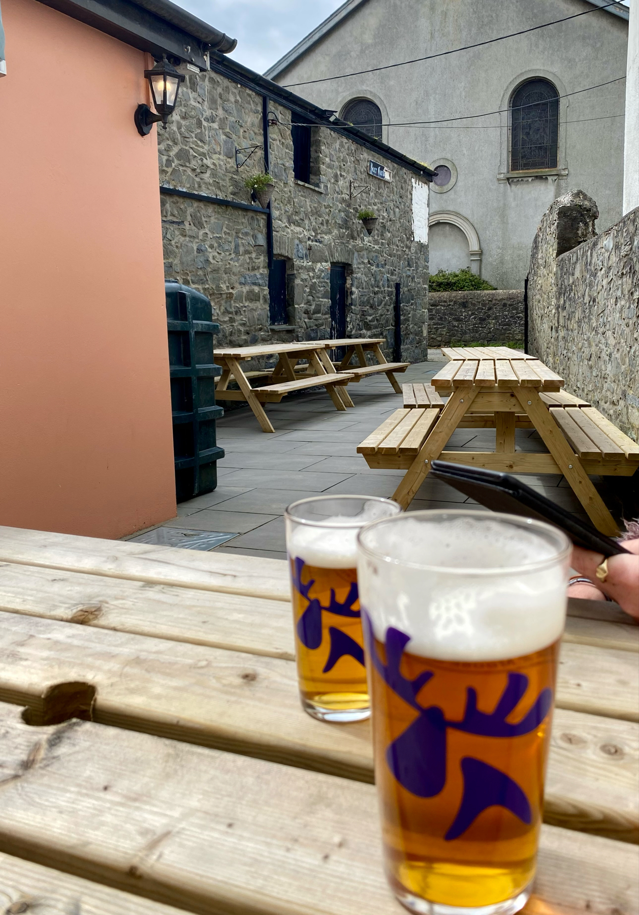 Two glasses of beer on a wooden table, with an outdoor seating area and stone buildings in the background.