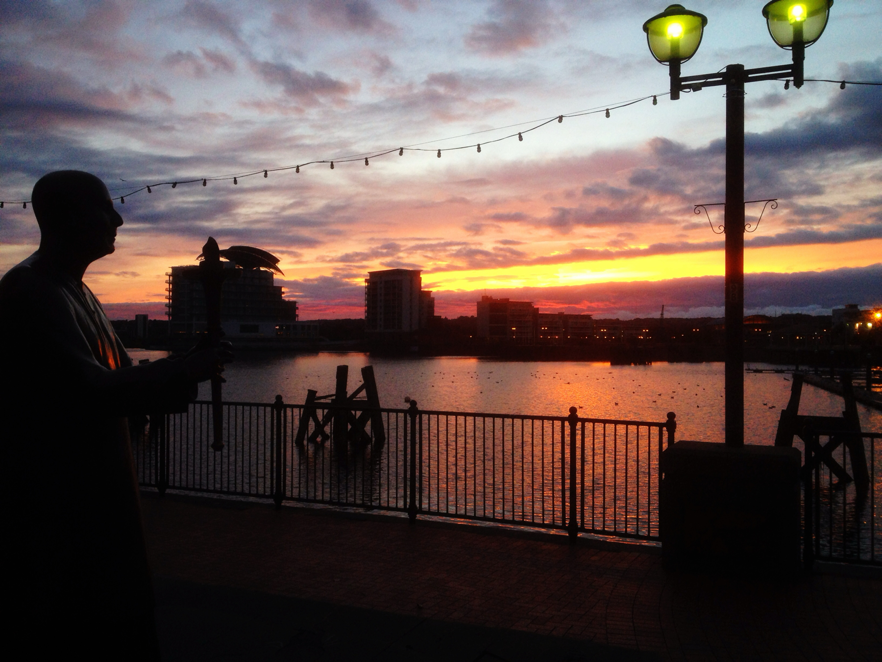 Silhouette of a statue by a waterfront at sunset, with street lights, buildings in the background, and a vividly colored sky.