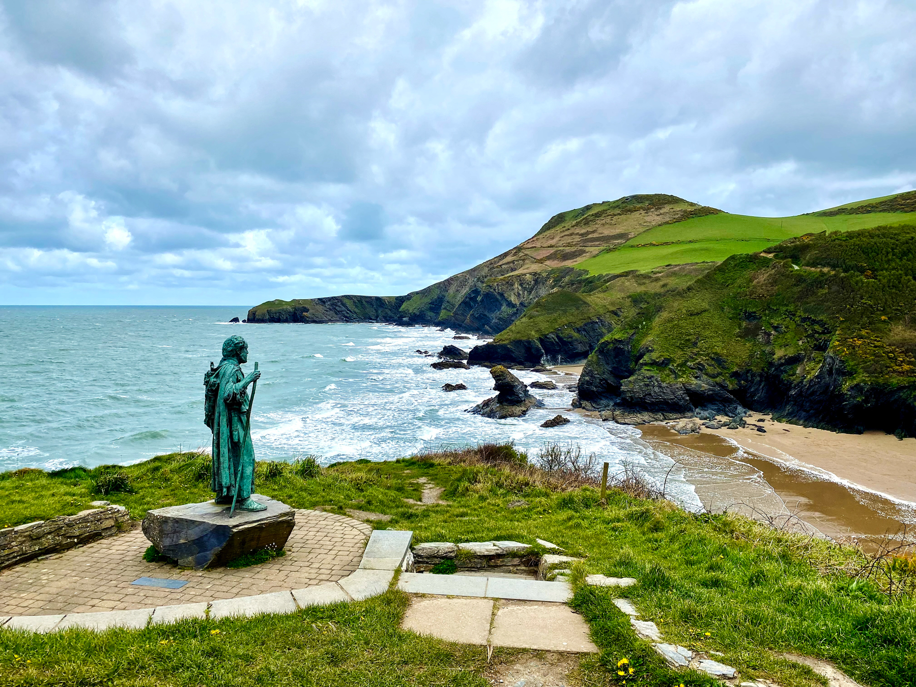 A bronze statue of a figure looking out to sea, standing on a cliff overlooking a rugged coastline with a sandy beach, rocky outcrops, and rolling green hills under a cloudy sky.