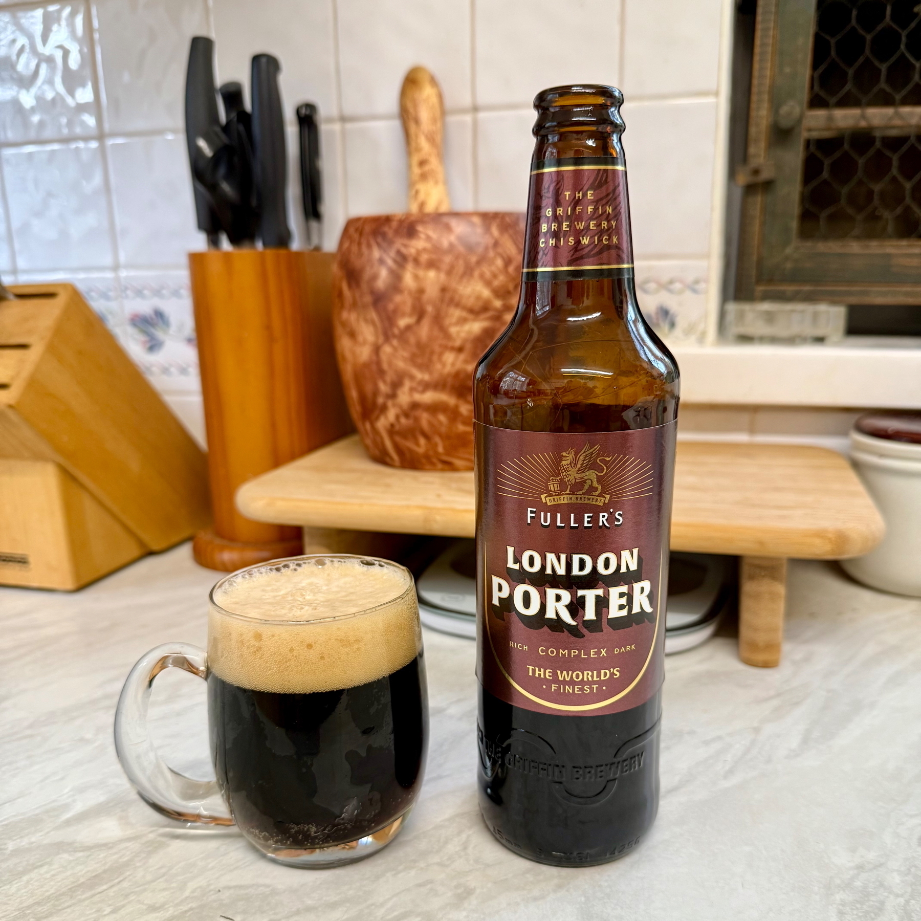 Bottle and glass of dark beer - Fullers London Porter - on a kitchen worktop. Knife block, chopping board and wooden pestle and mortar in the background.