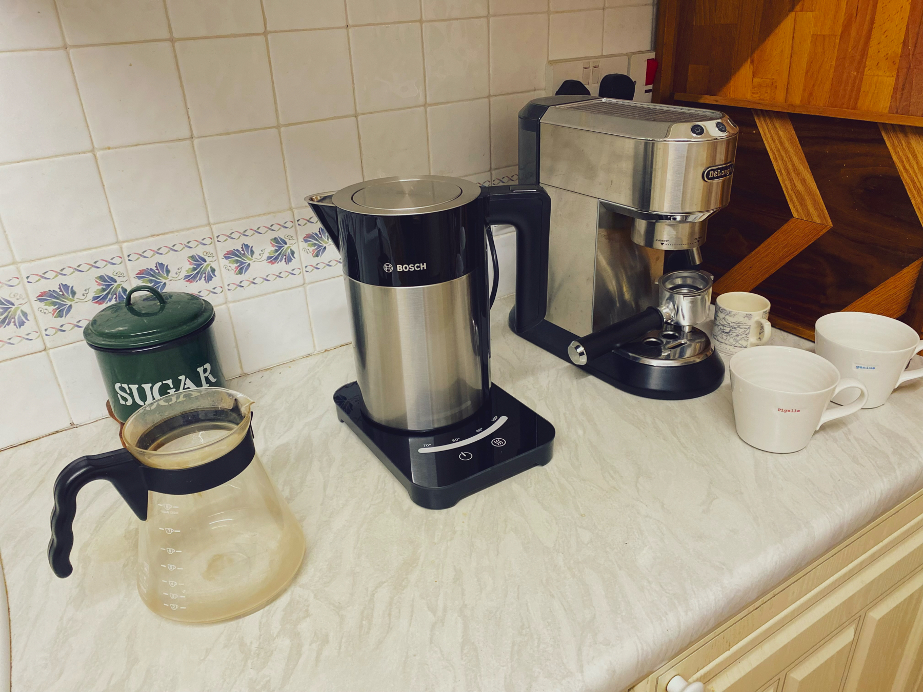 A kitchen countertop with a coffee-making setup including a kettle, an espresso machine, several mugs, and a sugar container.
