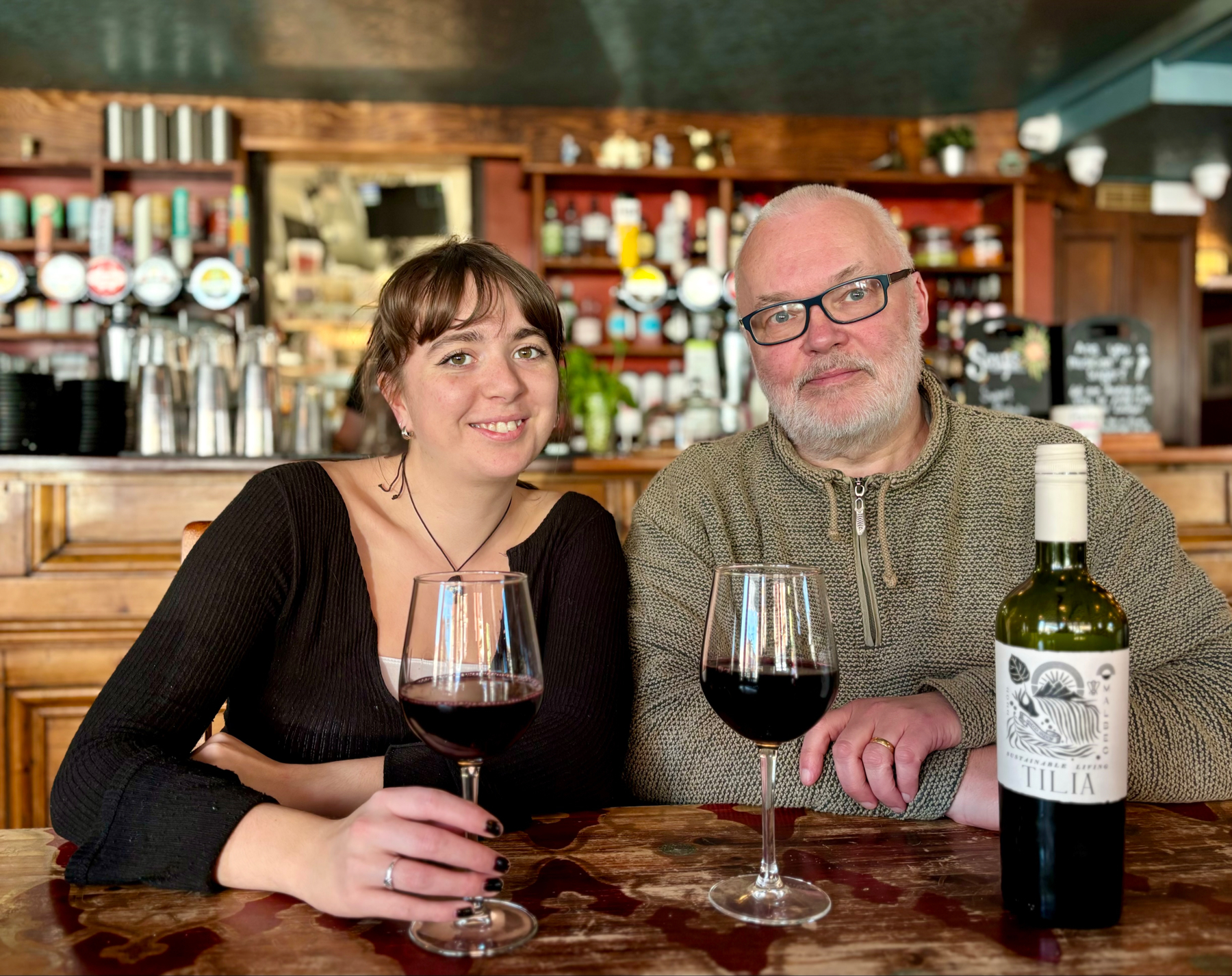 Two people sitting at a bar with glasses of red wine and a bottle of Tilia wine on the table.