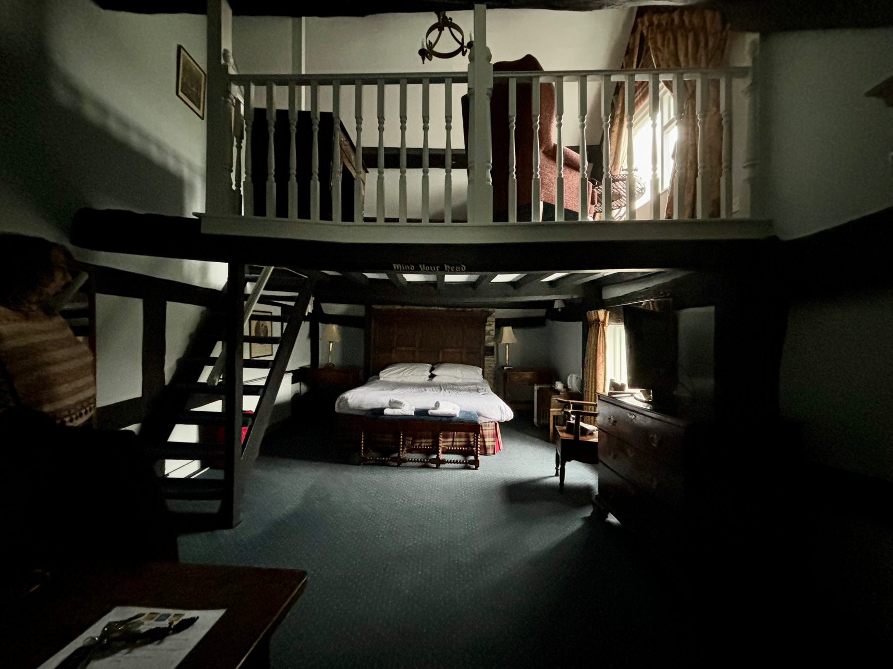 A dimly lit room featuring a traditional wooden loft with a ladder, a bed on the lower level, antique furniture including a dresser with a mirror, and a wicker trunk at the foot of the bed. Light filters in through a window