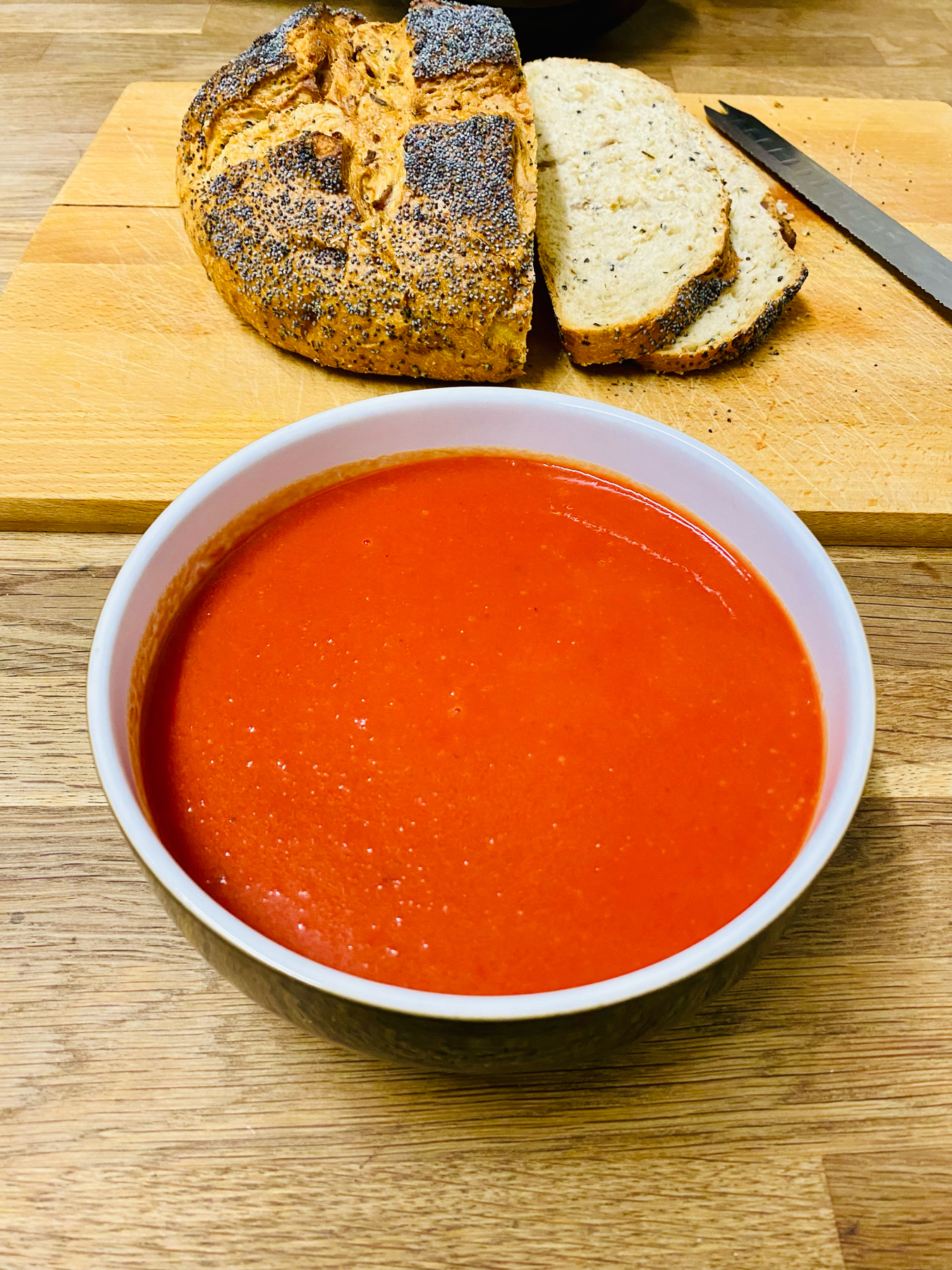 A bowl of soup with sliced multigrain bread and a whole loaf topped with poppy seeds on a wooden cutting board next to a knife.