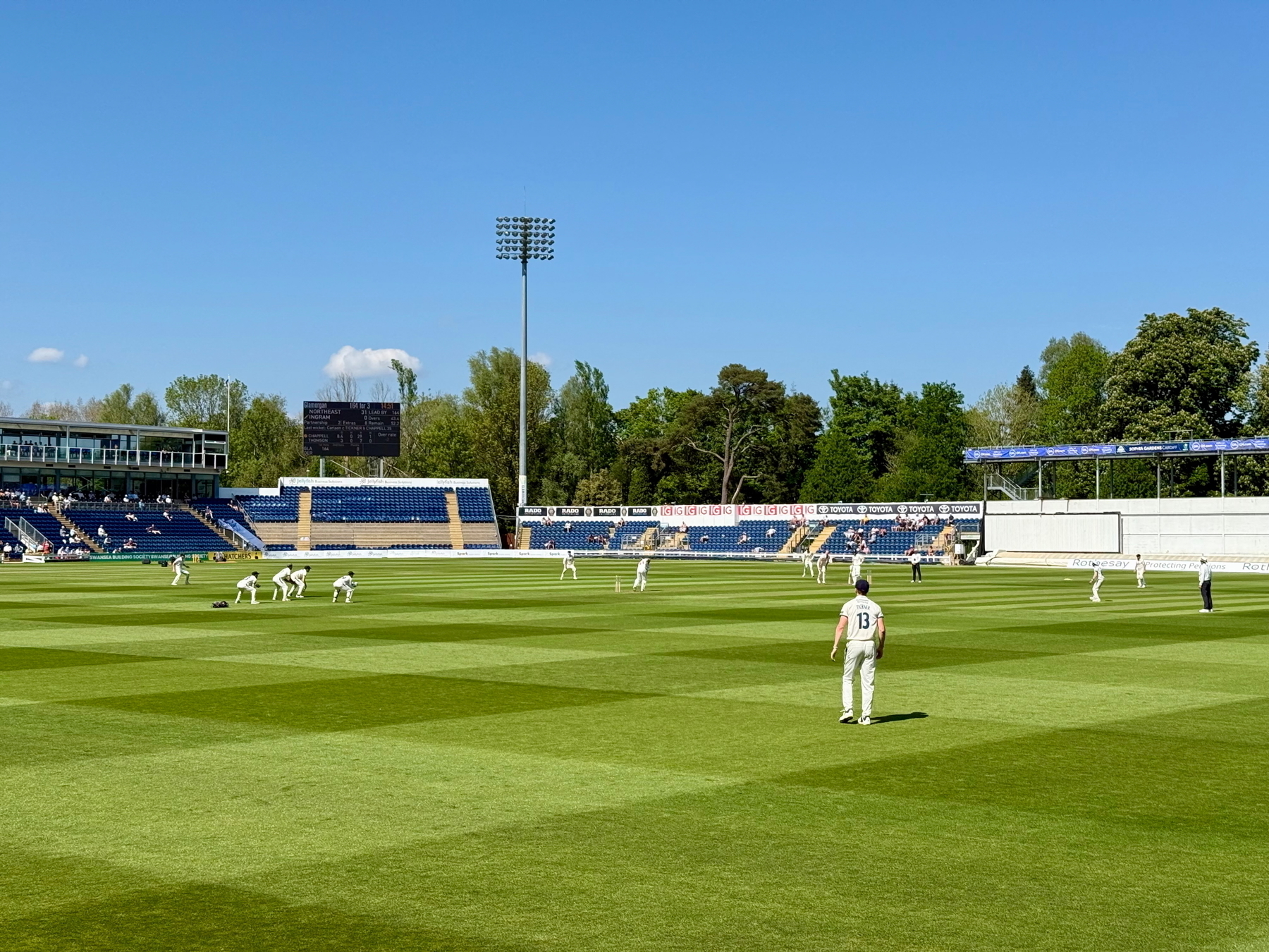 Auto-generated description: A cricket match is taking place on a sunny day with players on the field and spectators in the stands.