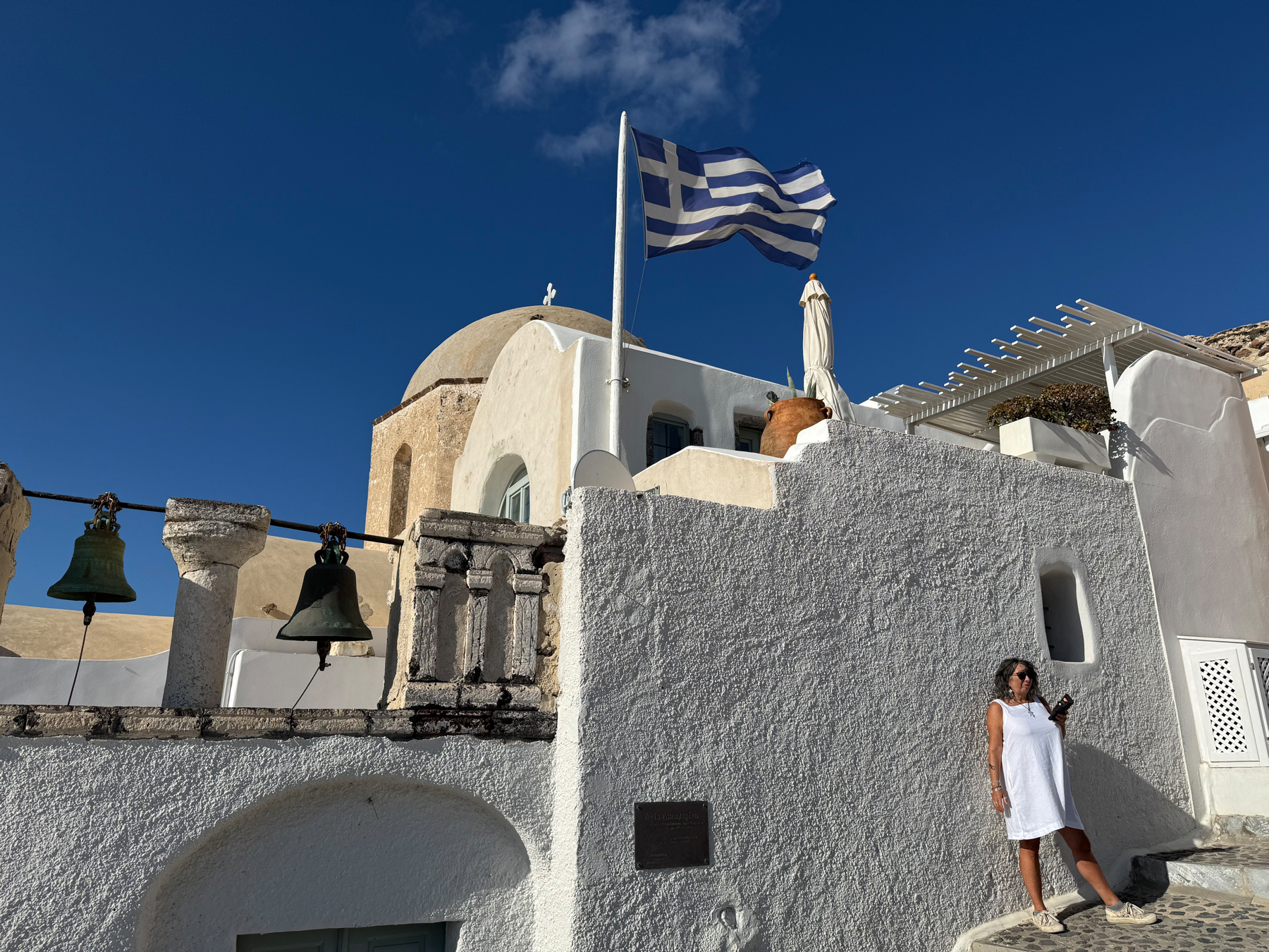Auto-generated description: A woman in a white dress stands on a whitewashed building with the Greek flag waving above and a deep blue sky in the background.