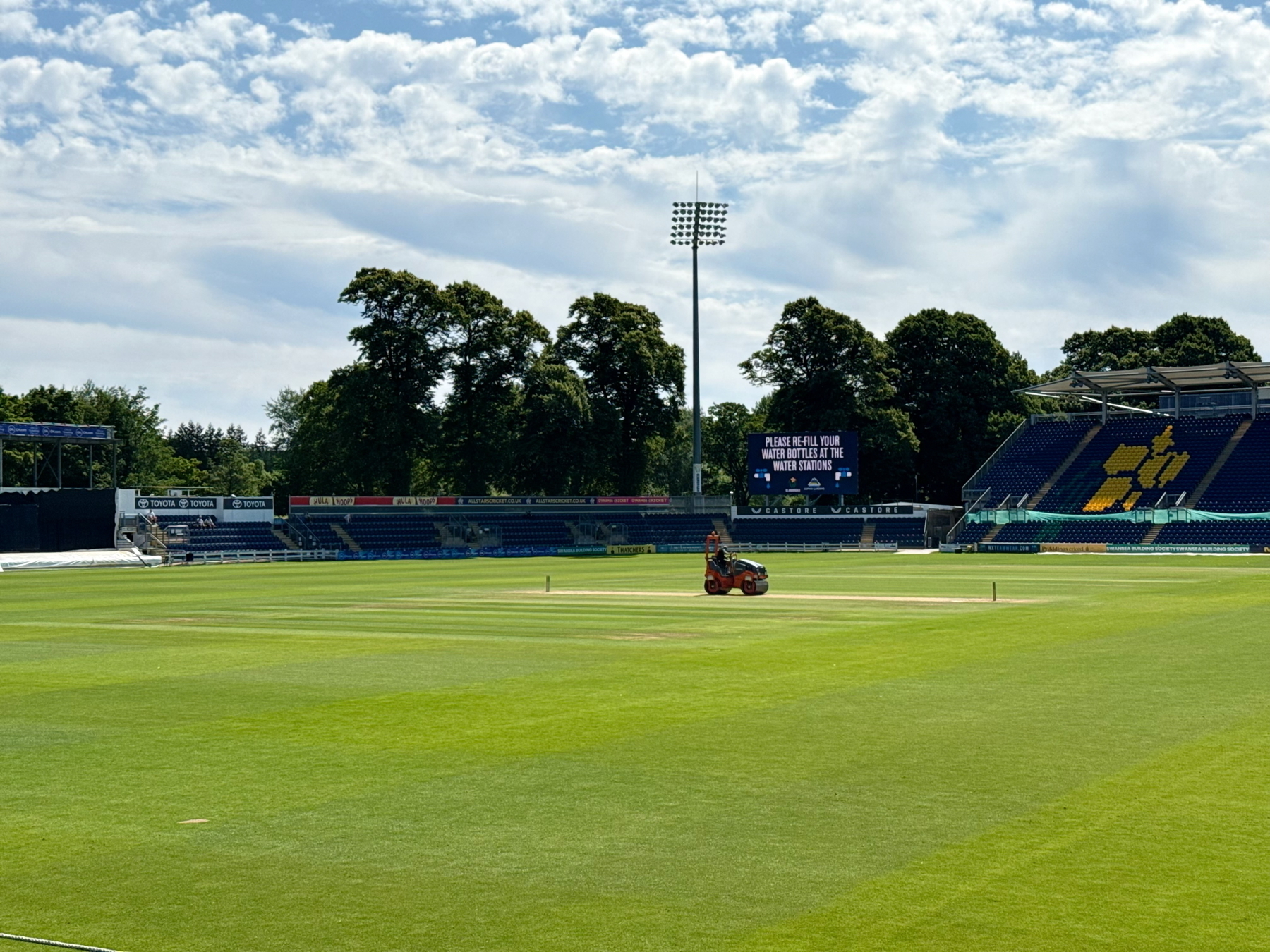 Auto-generated description: A person rides a lawnmower on a cricket field under a partly cloudy sky.