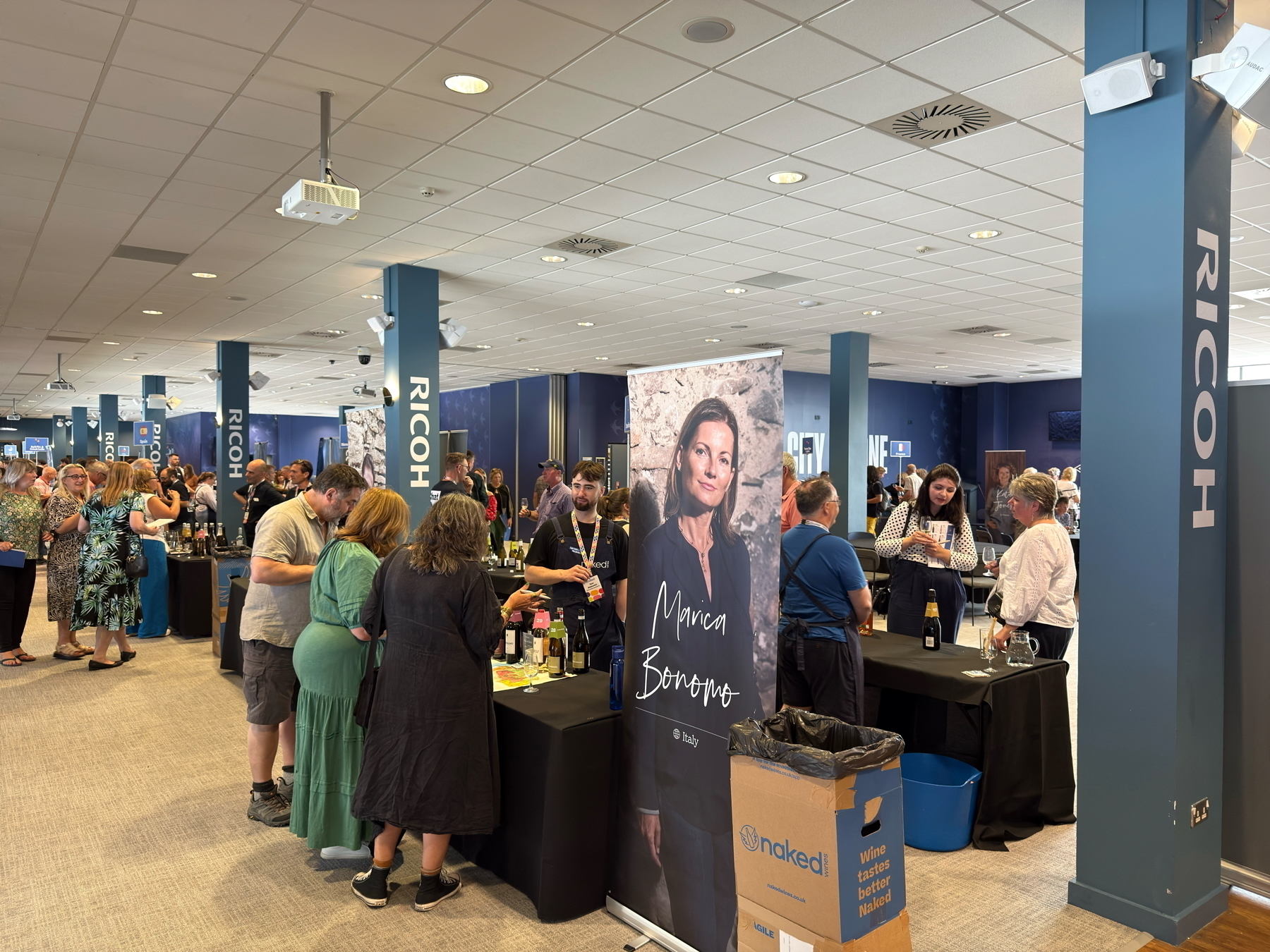 Auto-generated description: People are gathered in a spacious indoor setting with various booths and standing displays, including a prominent banner featuring a person named Mirca Bornova.