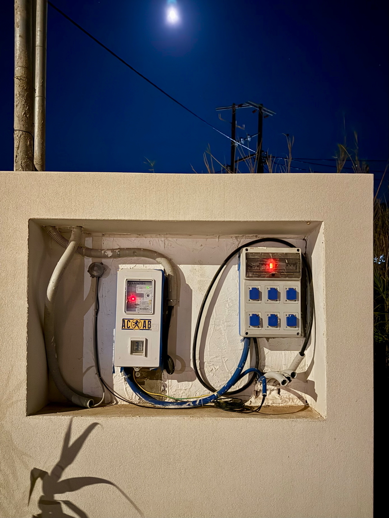 Auto-generated description: Two electrical boxes with wires are mounted on a white wall under a night sky with a visible moon.