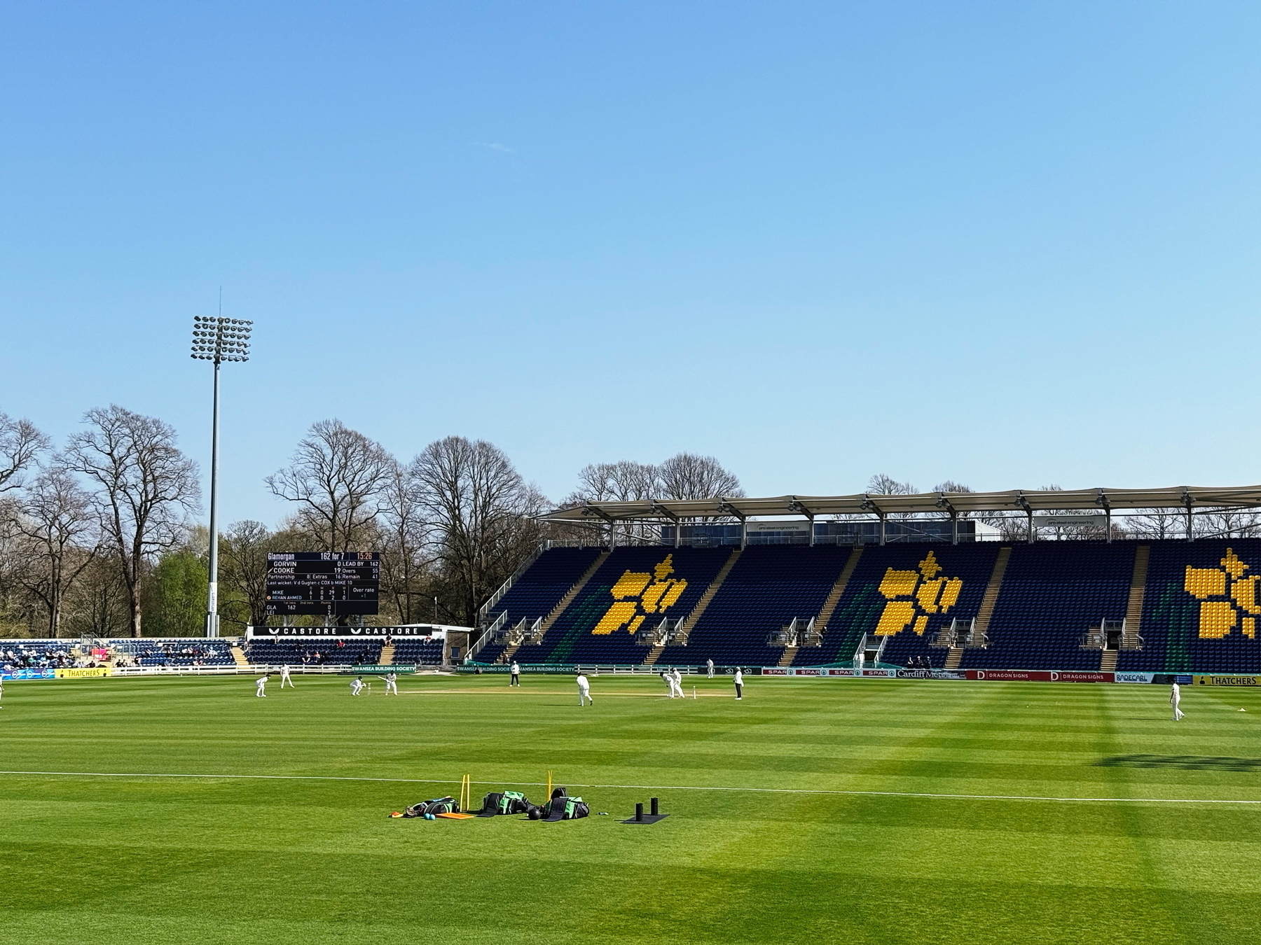Auto-generated description: A cricket match is taking place on a sunny day in a stadium with blue seating and a partially filled crowd.