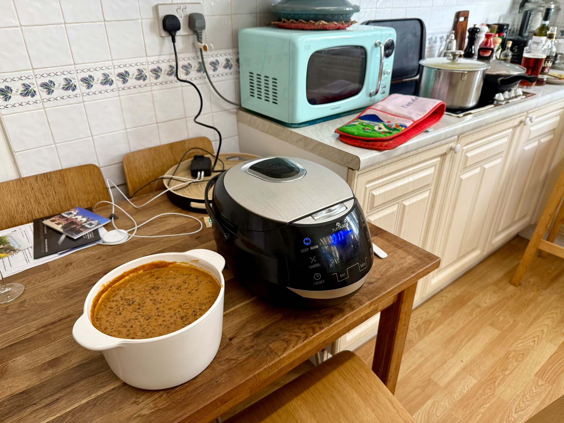 Auto-generated description: A kitchen scene features a wooden table with a pot of dal and a rice cooker, set against a background of a microwave, pots, and cooking utensils on the counter.