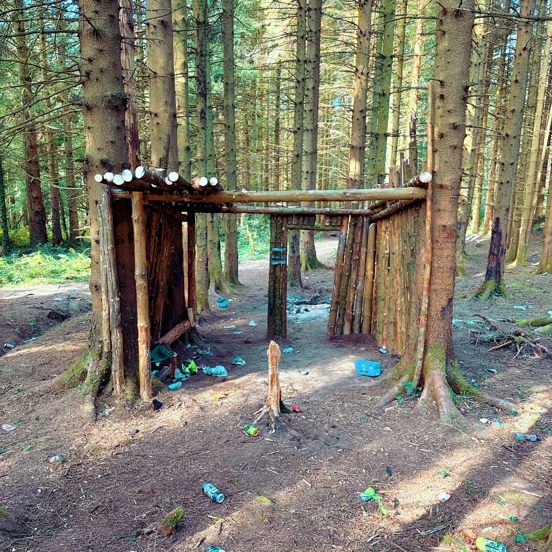 A wooden shelter built with logs is situated in a forest. The ground around the shelter is littered with trash, including plastic bottles and cans. Tall trees surround the area, and sunlight filters through the branches.