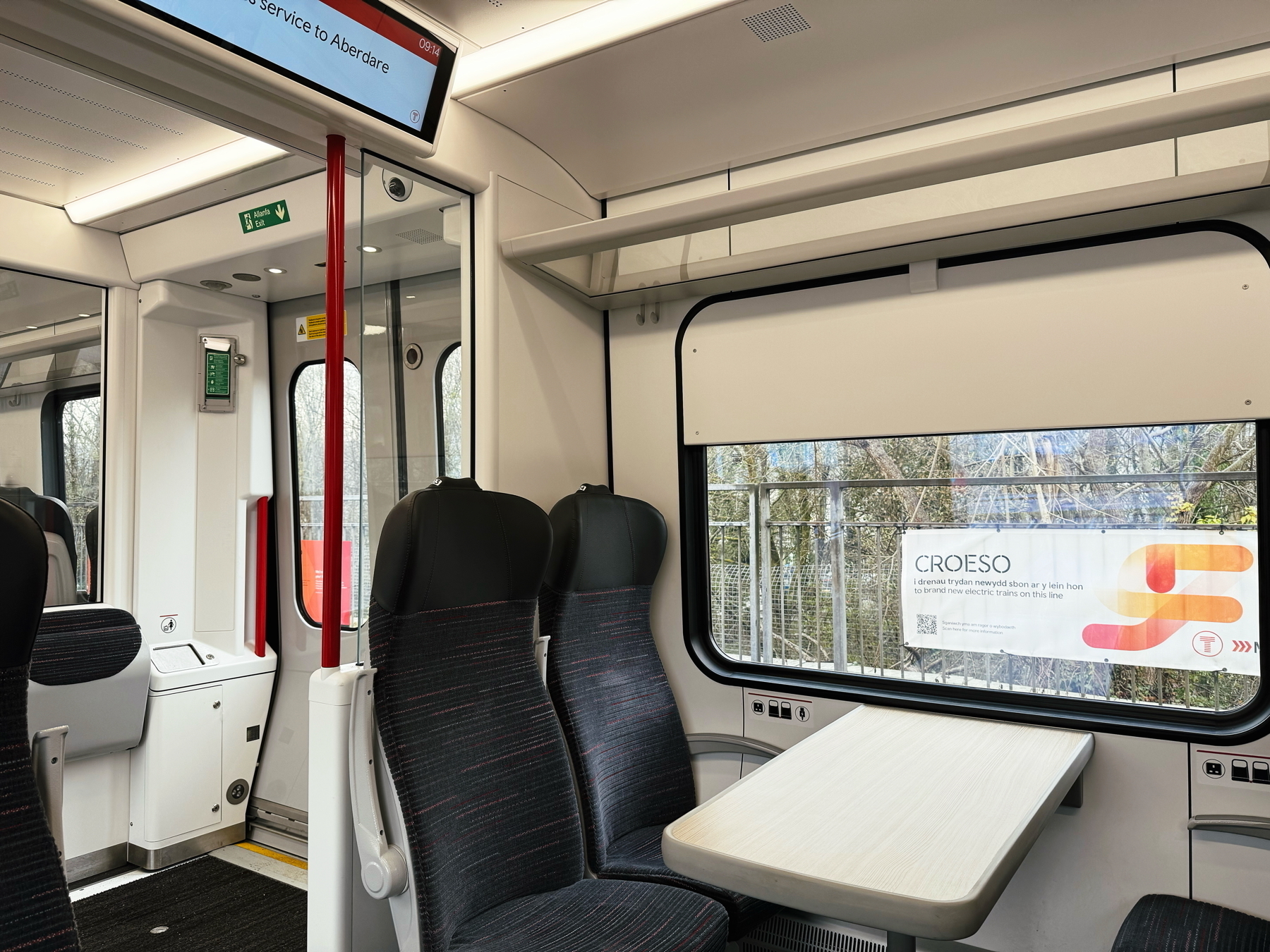 Auto-generated description: A modern train interior features seating around a table, with large windows and electronic signage displaying travel information.
