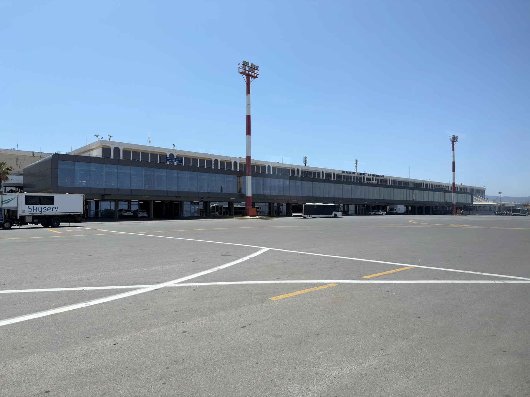 Auto-generated description: A large, modern airport terminal with several vehicles in front and clear blue skies above.
