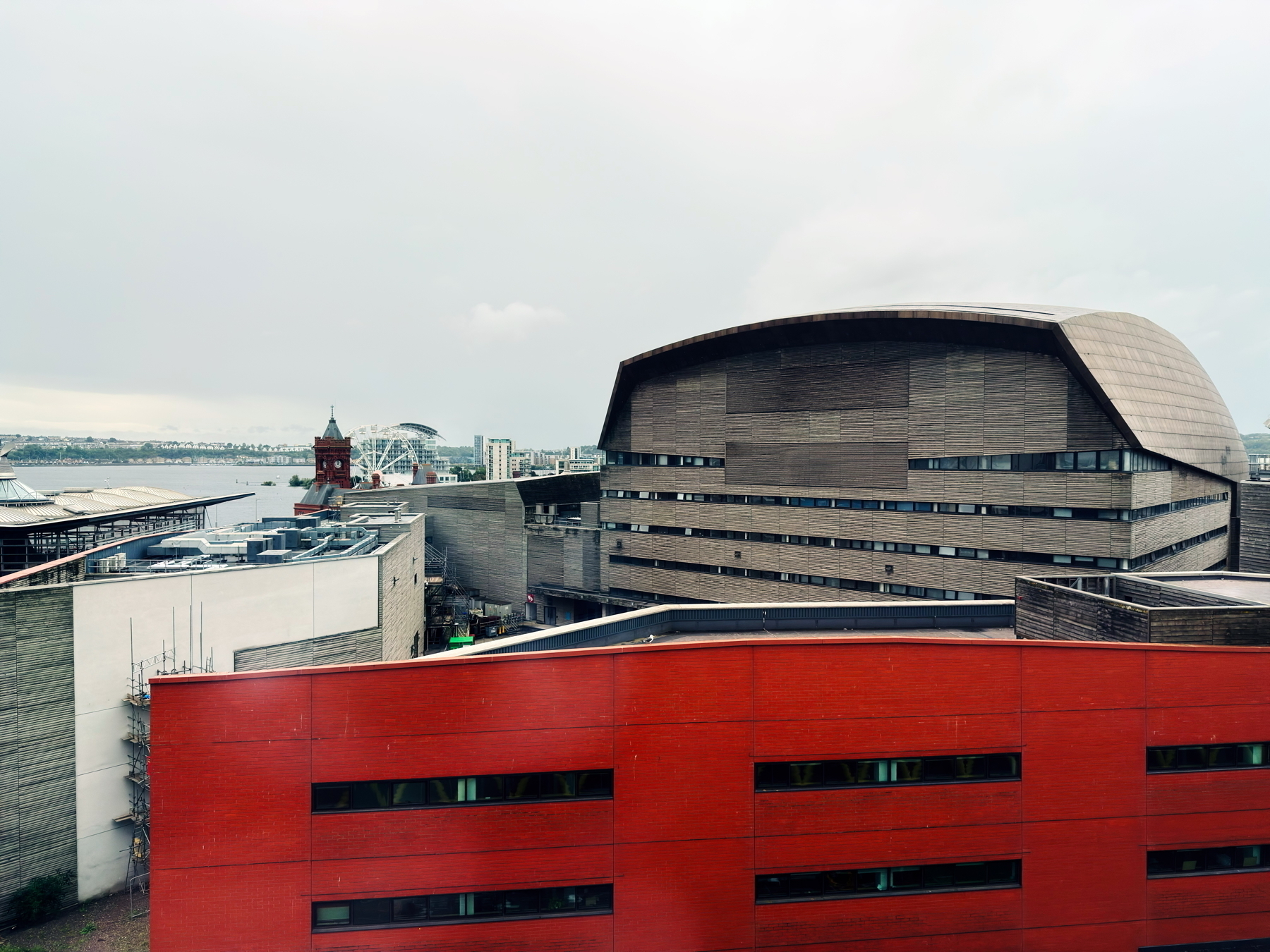 The image shows a large modern building with a distinctive, curved roof, likely an arts or cultural center, set against a cloudy sky. In the foreground, there is a red brick building. In the background, a river with a bridge and Ferris wheel.