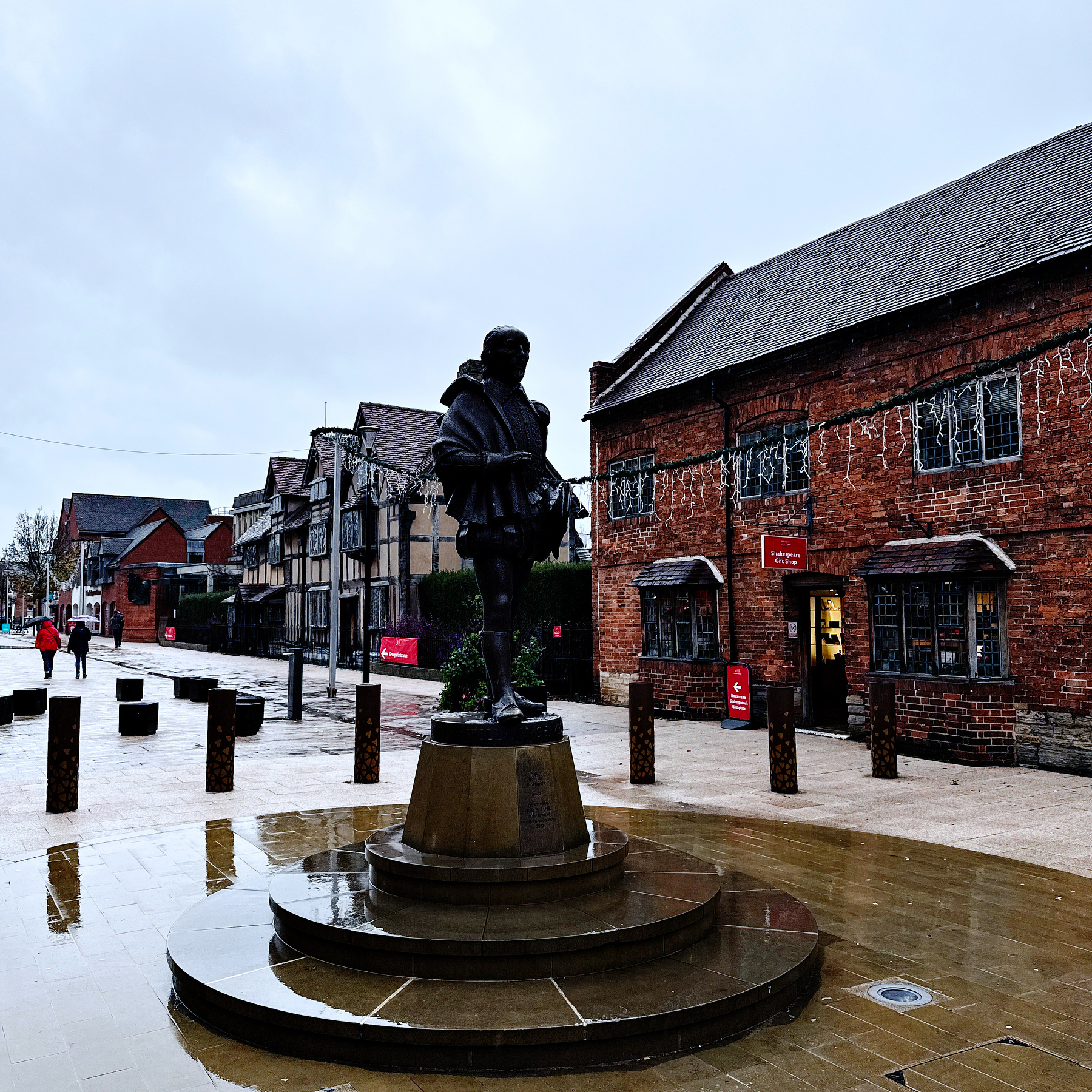 Auto-generated description: A statue stands on a pedestal in a paved outdoor area surrounded by historic brick buildings and festive lights.