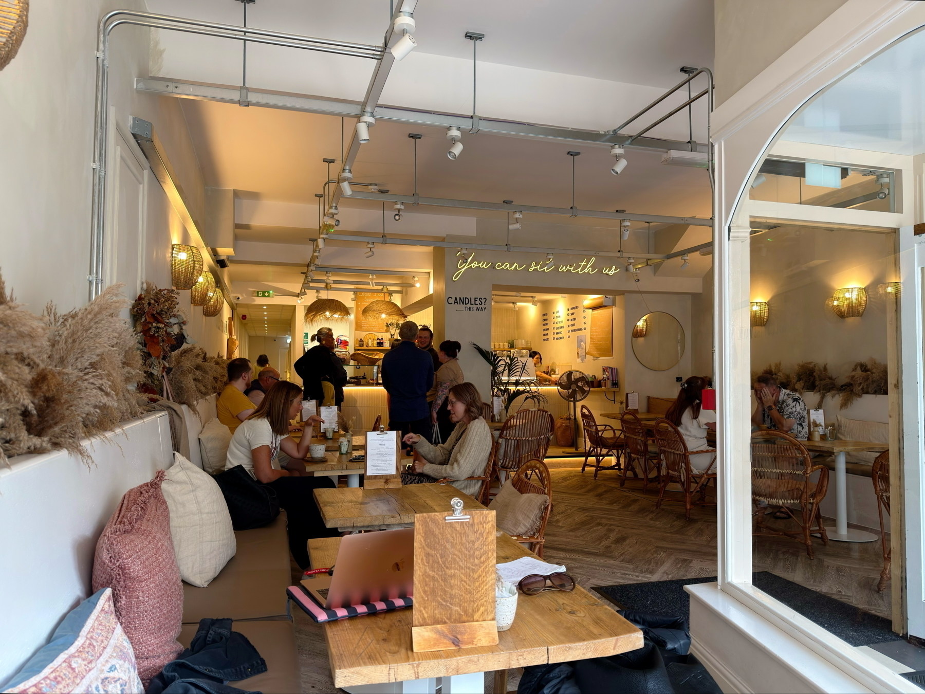 Auto-generated description: A cozy café interior features customers sitting at wooden tables amidst warm lighting and decorative pampas grass.