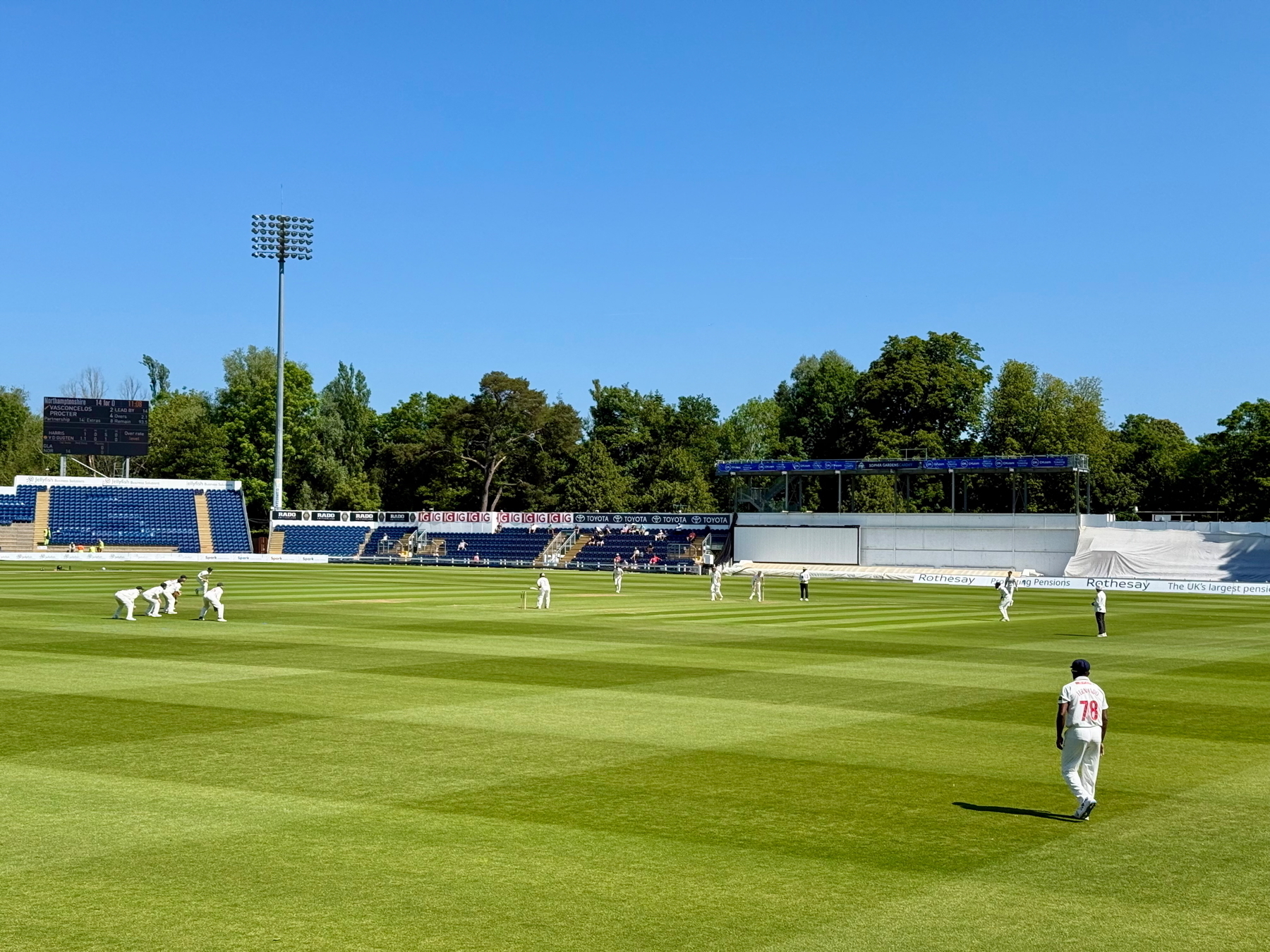 Auto-generated description: A cricket match is taking place on a sunlit ground with players in white uniforms and spectators in the stands.