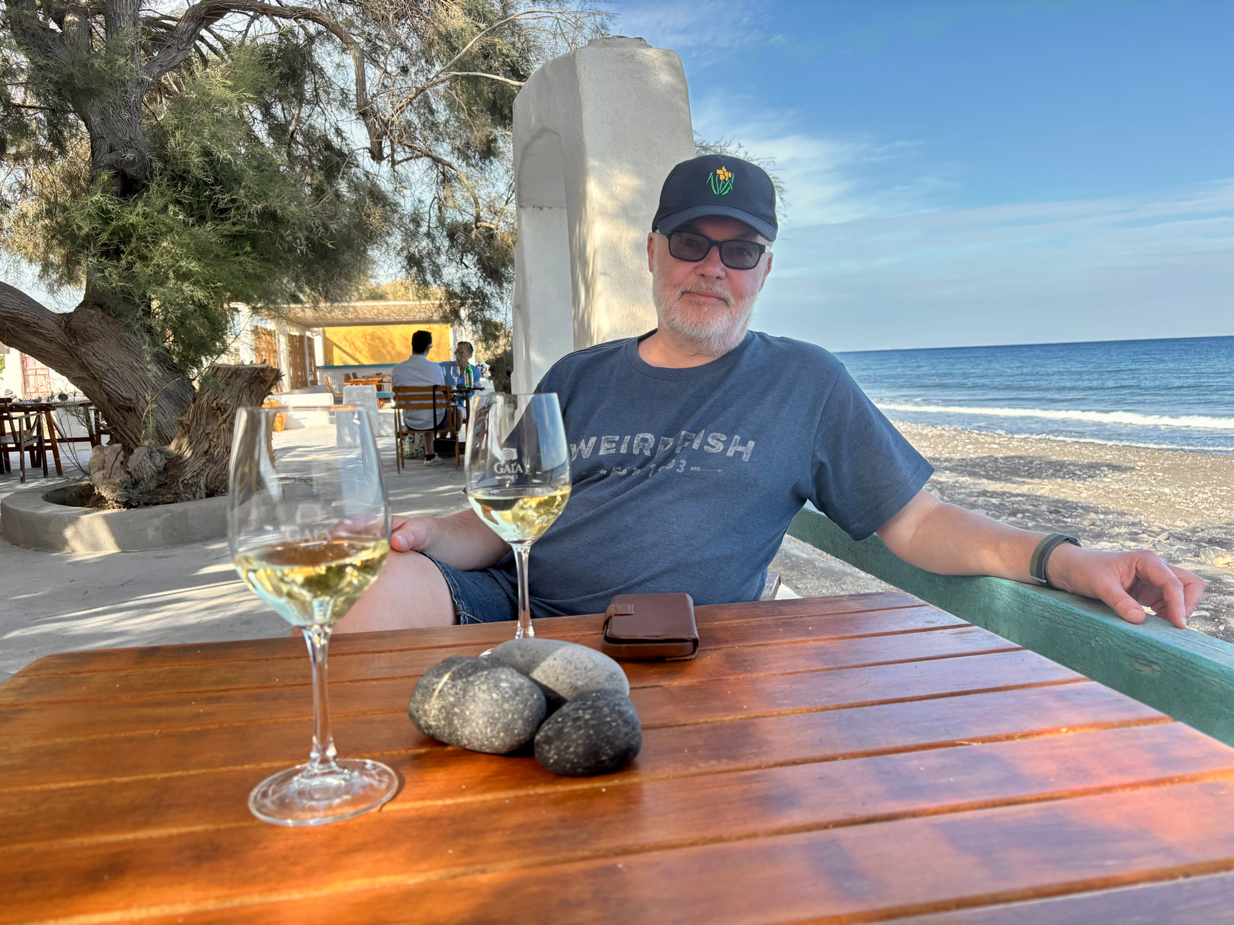 Auto-generated description: A man wearing sunglasses and a cap relaxes at an outdoor table by the beach with two glasses of white wine and some stones on the table.