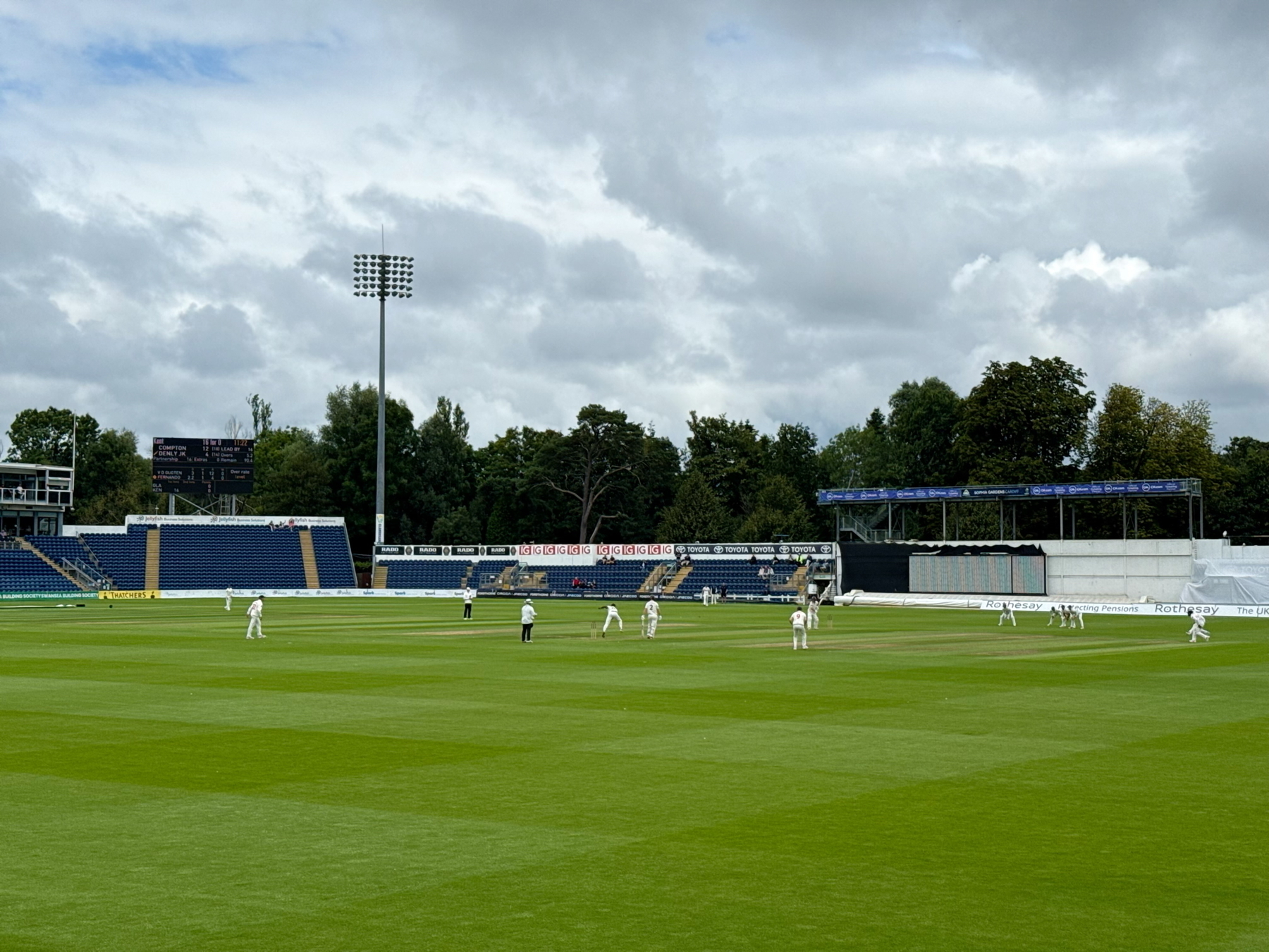 Auto-generated description: A cricket match is taking place on a lush green field under a cloudy sky, with players in white uniforms scattered across the pitch.