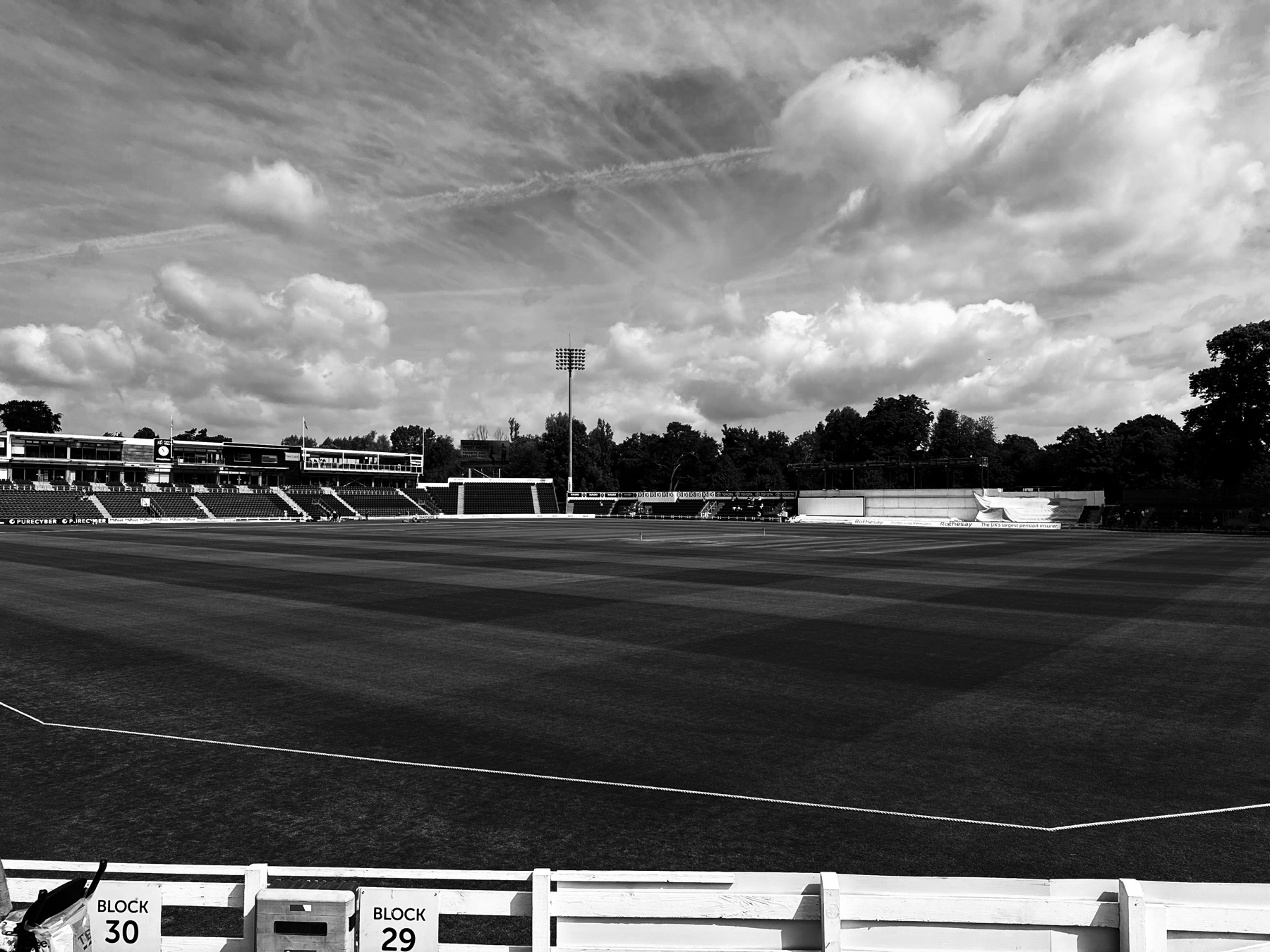 Auto-generated description: A cricket stadium with an expansive field and stands under a partly cloudy sky.