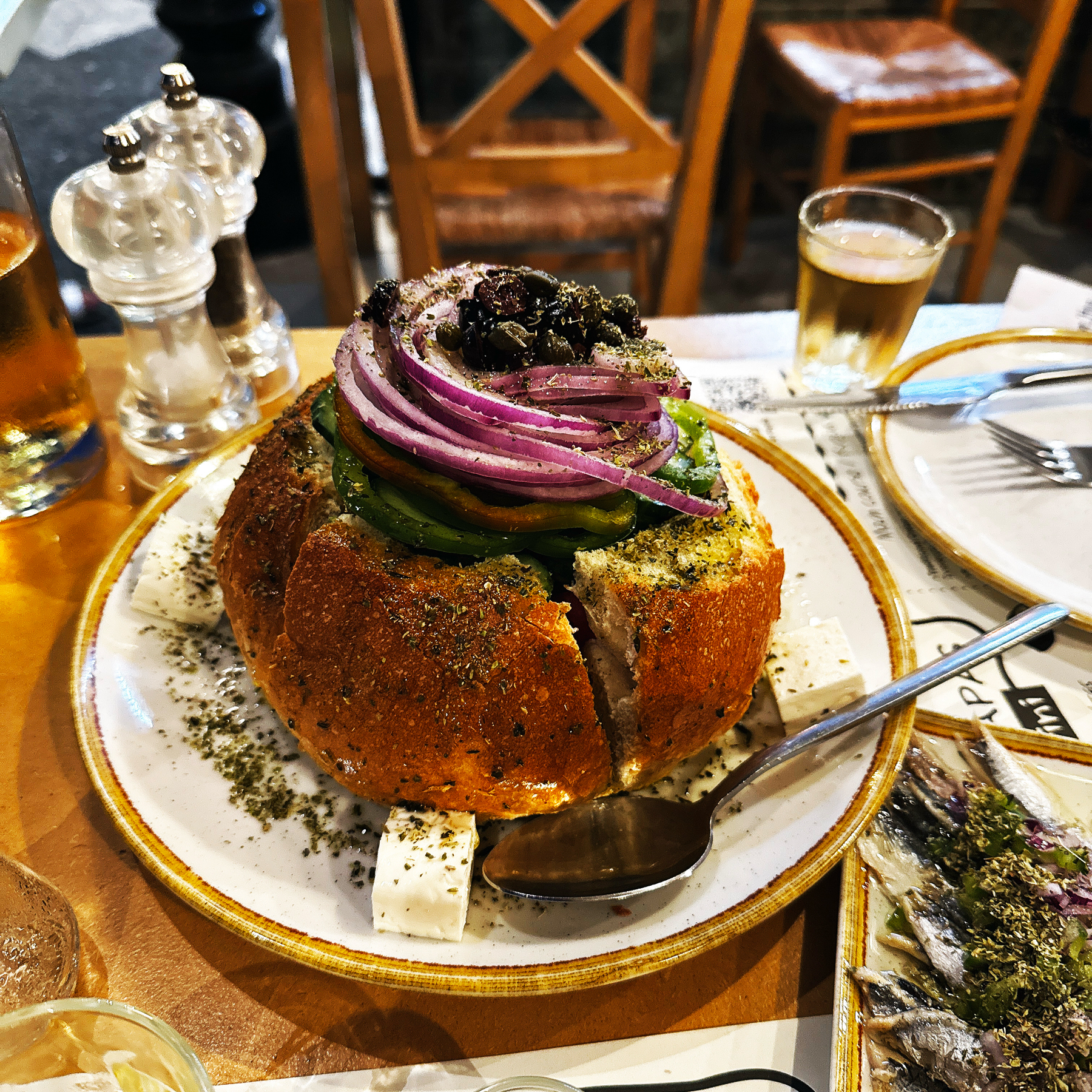 A bread bowl filled with salad topped with sliced onions and olives is surrounded by feta cheese and herbs on a plate set at a dining table.
