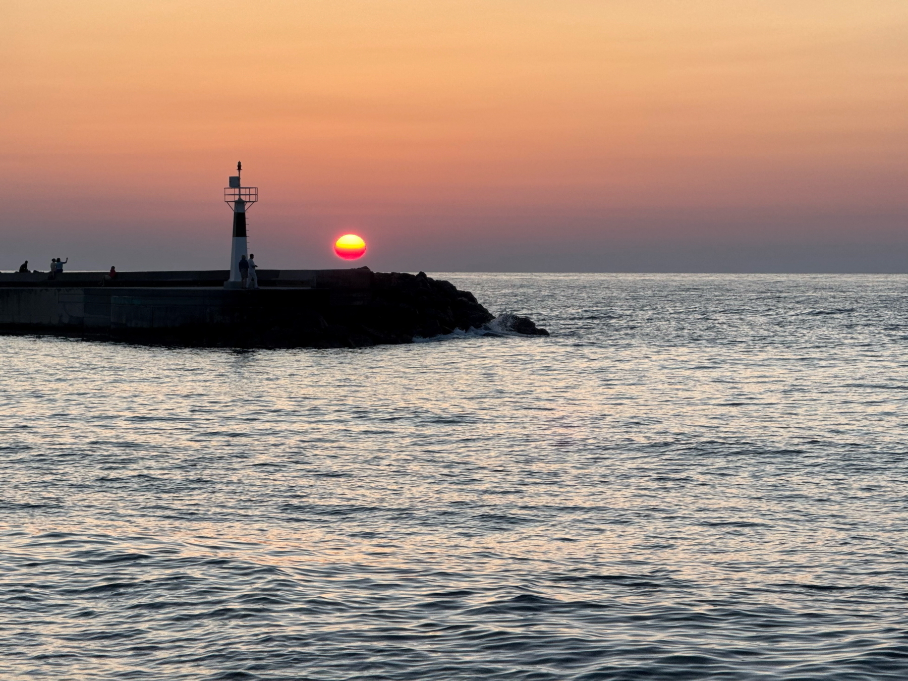 Auto-generated description: A serene sunset over the ocean with a lighthouse on a rocky pier.
