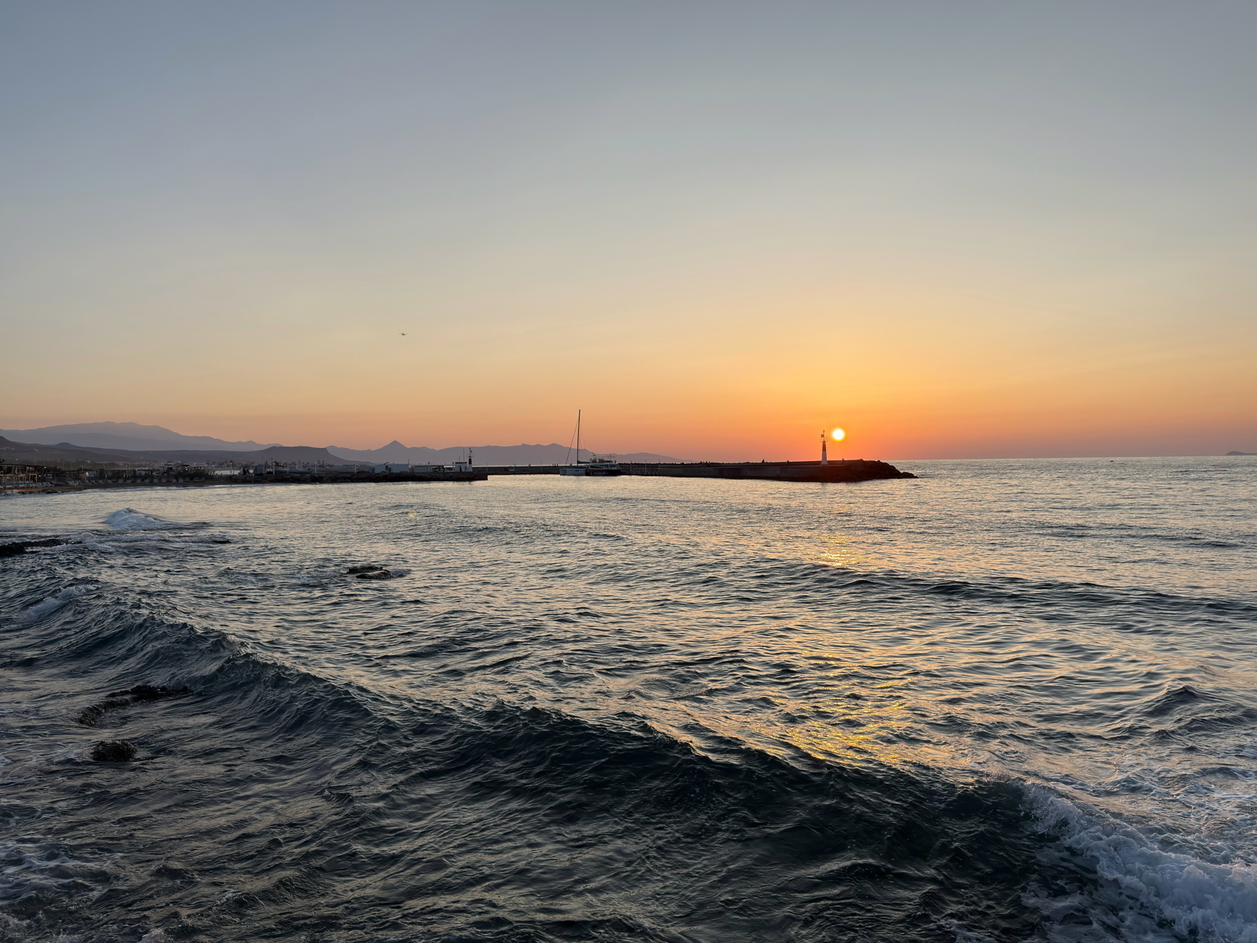 Auto-generated description: A tranquil sunset over the ocean with gentle waves and a distant sailboat near the horizon.