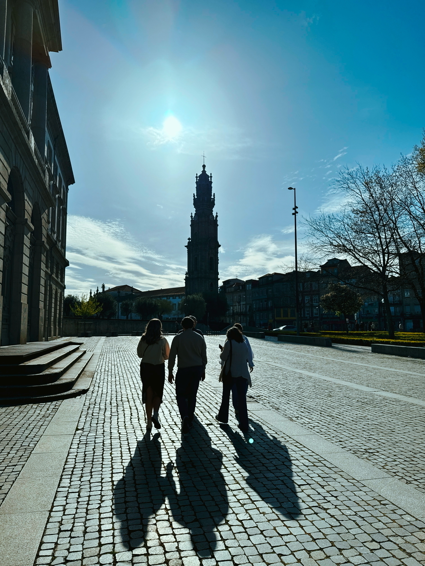 Auto-generated description: Three people walk along a cobblestone street, casting long shadows, with a tall tower and clear blue sky in the background.