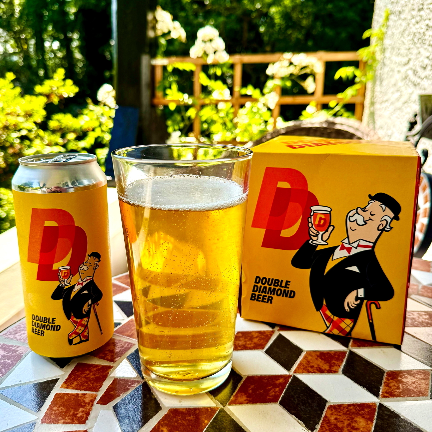 Auto-generated description: A can and box of Double Diamond Beer are on a patterned table next to a glass filled with beer, with outdoor greenery in the background.