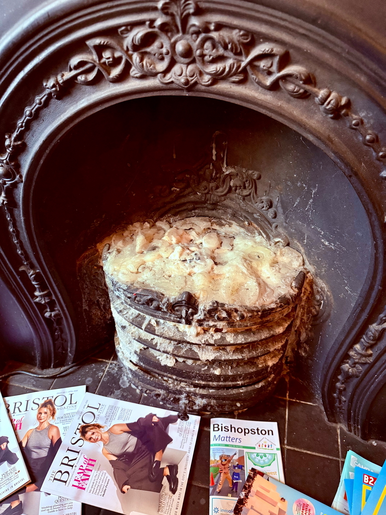 Auto-generated description: A vintage fireplace is stacked with wax-dripped, partially melted candles, surrounded by numerous colorful magazines on the floor.