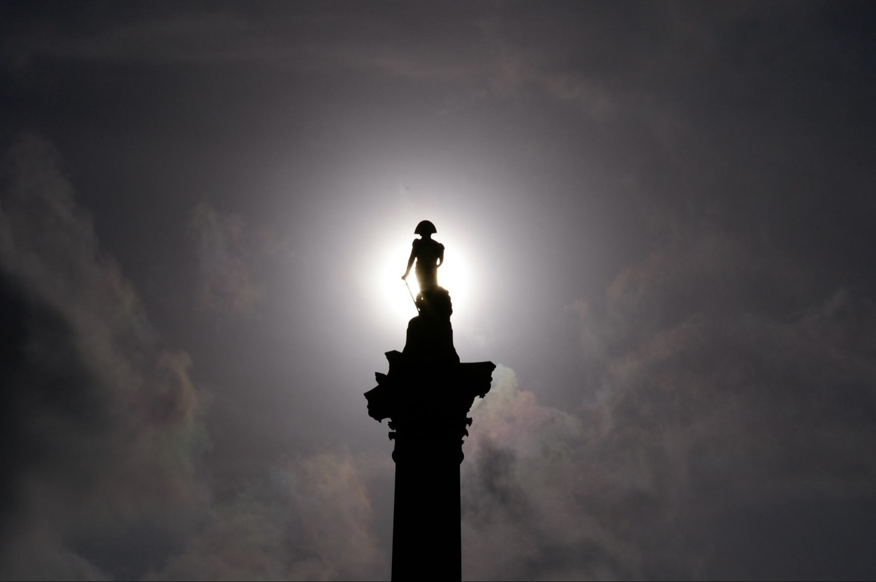 Auto-generated description: A silhouette of a statue stands atop a column against a dramatic sky with the sun creating a halo effect around it.