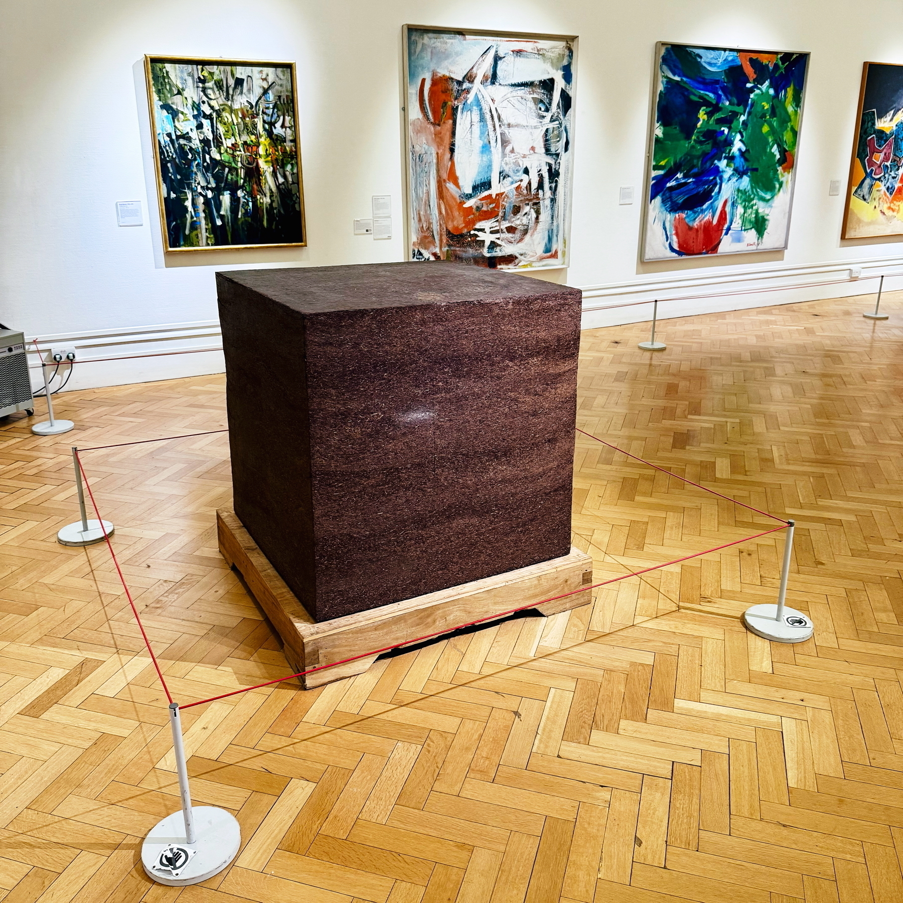 Auto-generated description: A large cubic sculpture is displayed in a gallery room with abstract paintings on the walls.