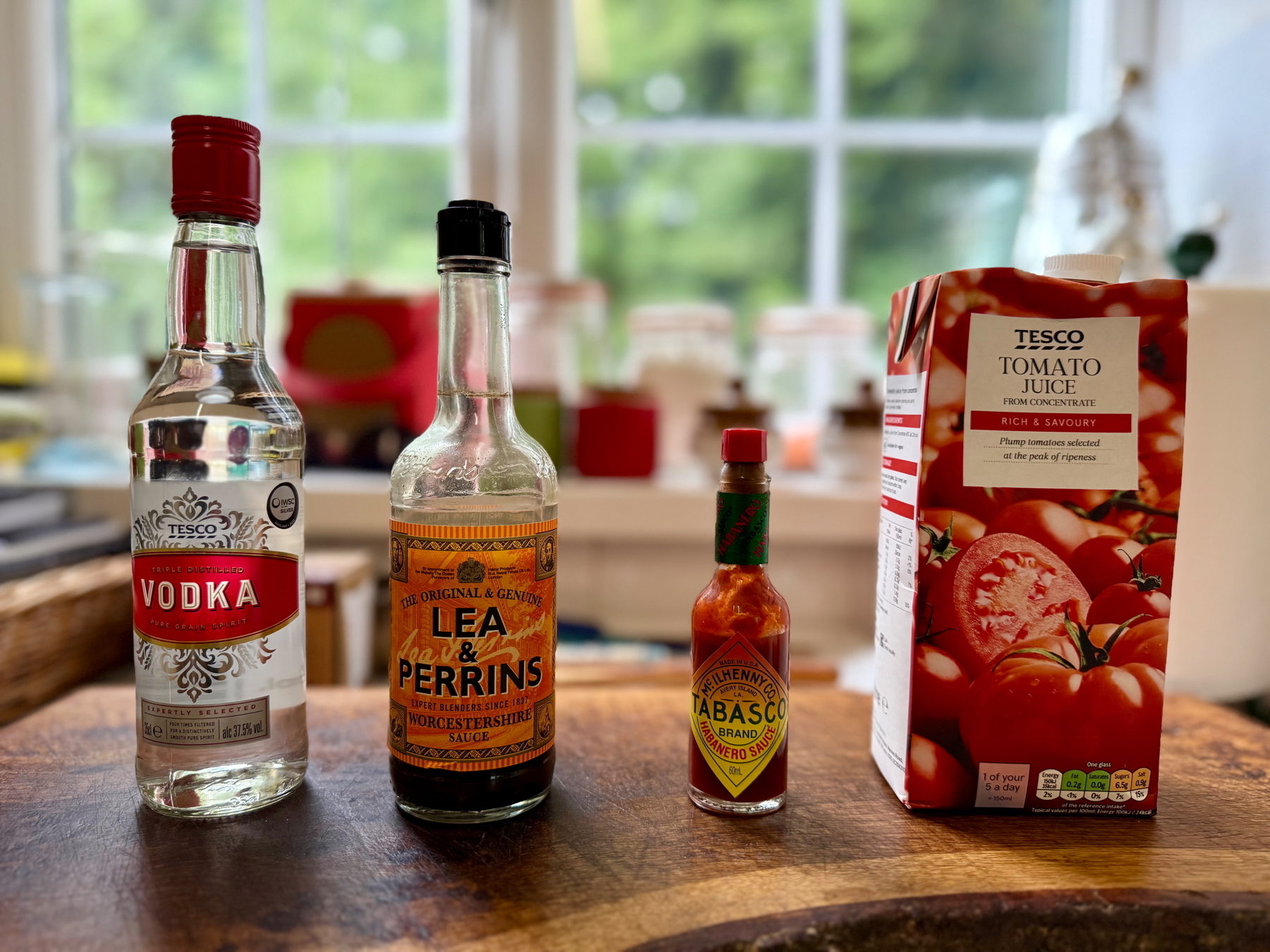 Auto-generated description: Bottles of vodka, Worcestershire sauce, Tabasco sauce, and a carton of tomato juice are lined up on a kitchen counter.
