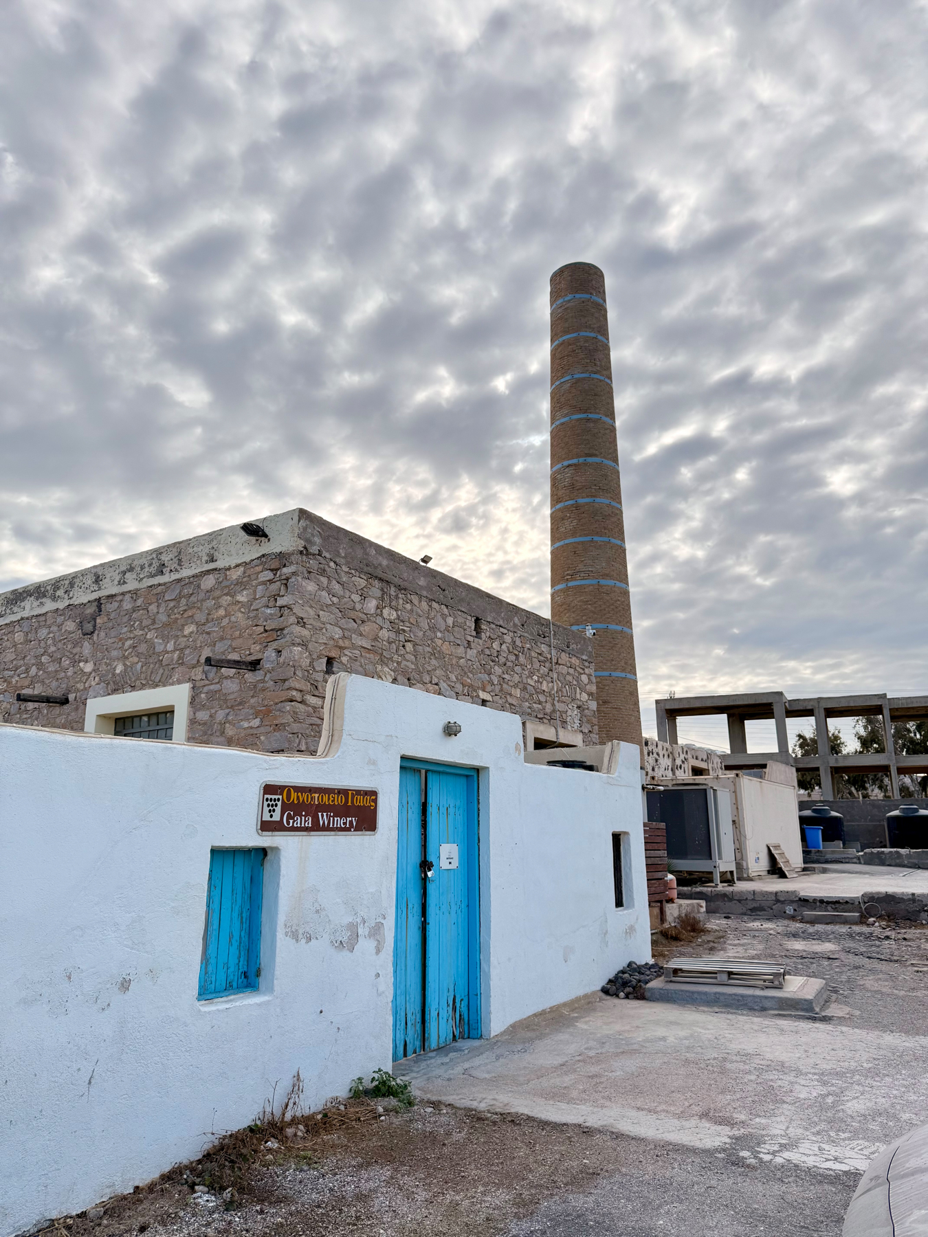 Auto-generated description: A rustic building with a blue door and a tall chimney stands under a cloudy sky, with a sign that reads Gaia Winery.