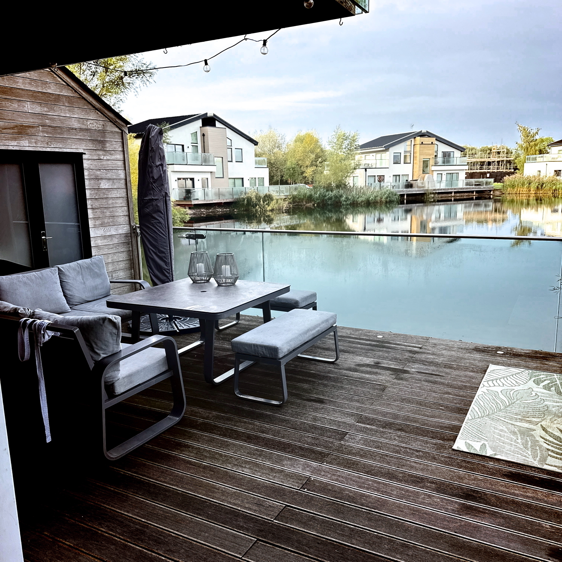 A modern lakeside deck with a wooden floor features a dining table set with cushioned chairs and benches. Two lanterns sit on the table. The deck overlooks a peaceful lake surrounded by contemporary houses with large windows.