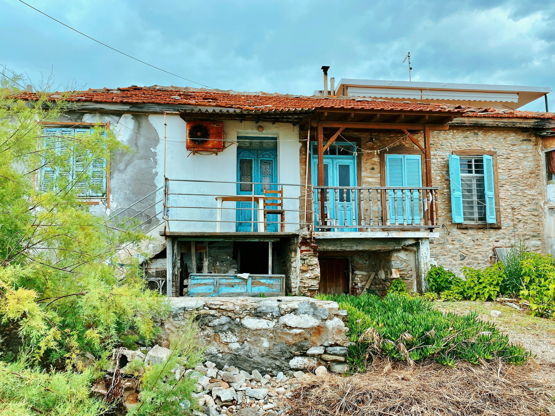 Auto-generated description: A rustic, weathered house with blue doors and shutters stands amid greenery under a cloudy sky.