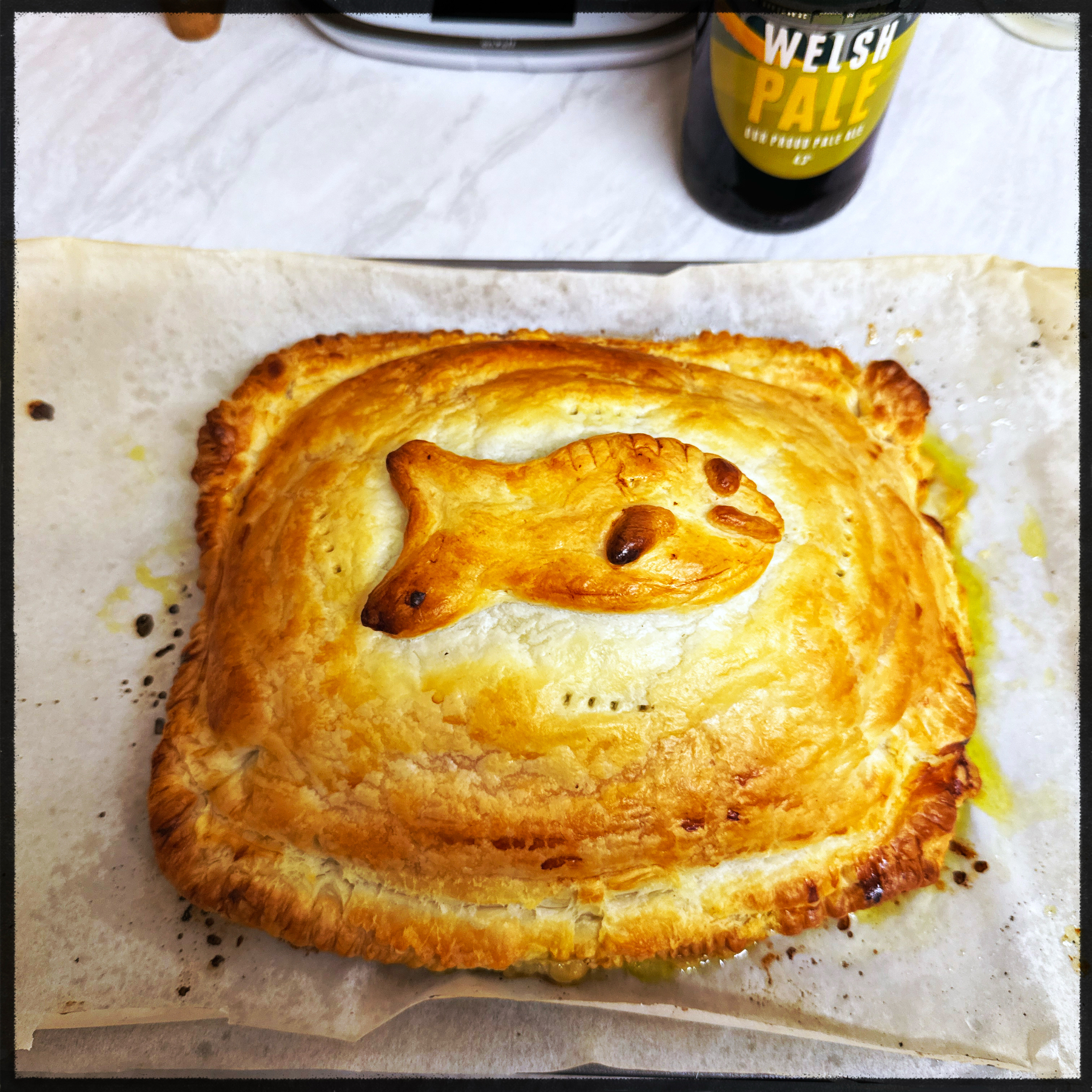 Auto-generated description: A baked pastry shaped like a fish rests on parchment paper, with a bottle of Welsh pale ale in the background.