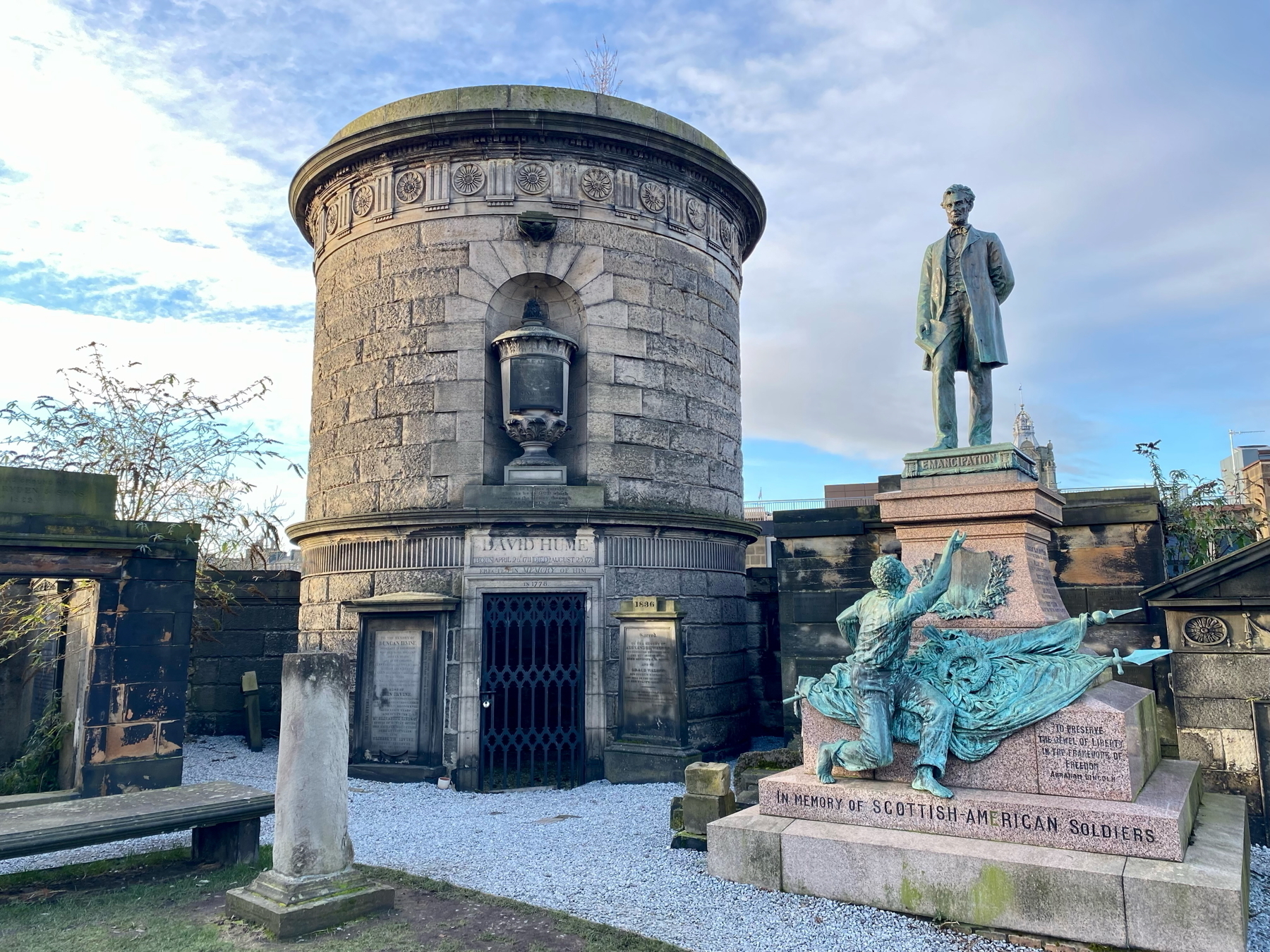 Auto-generated description: A historical stone monument and a statue dedicated to Scottish-American soldiers are set against a partly cloudy sky.