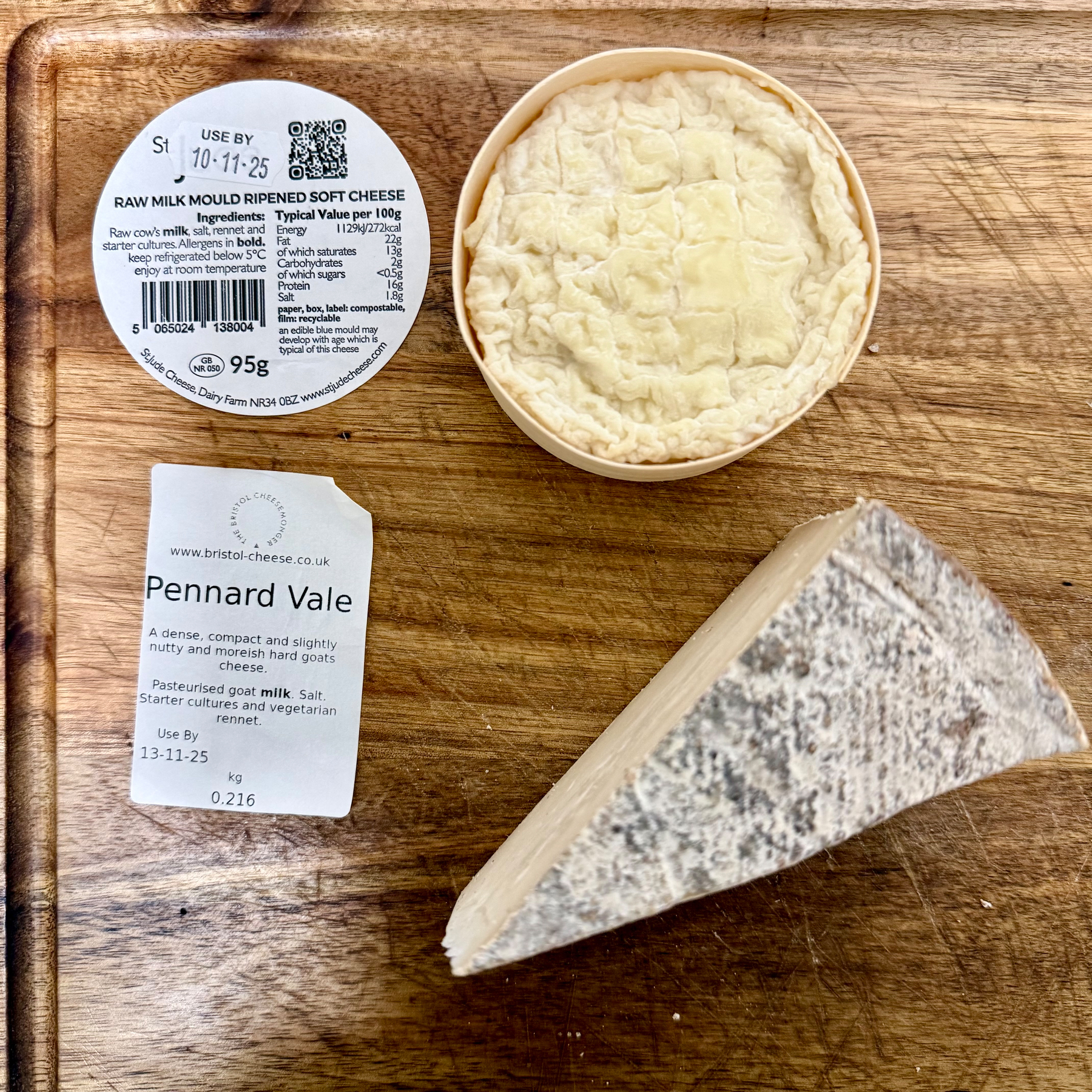Auto-generated description: A wedge of cheese and a round of mold-ripened cheese are displayed on a wooden board, accompanied by labels detailing their names and ingredients.