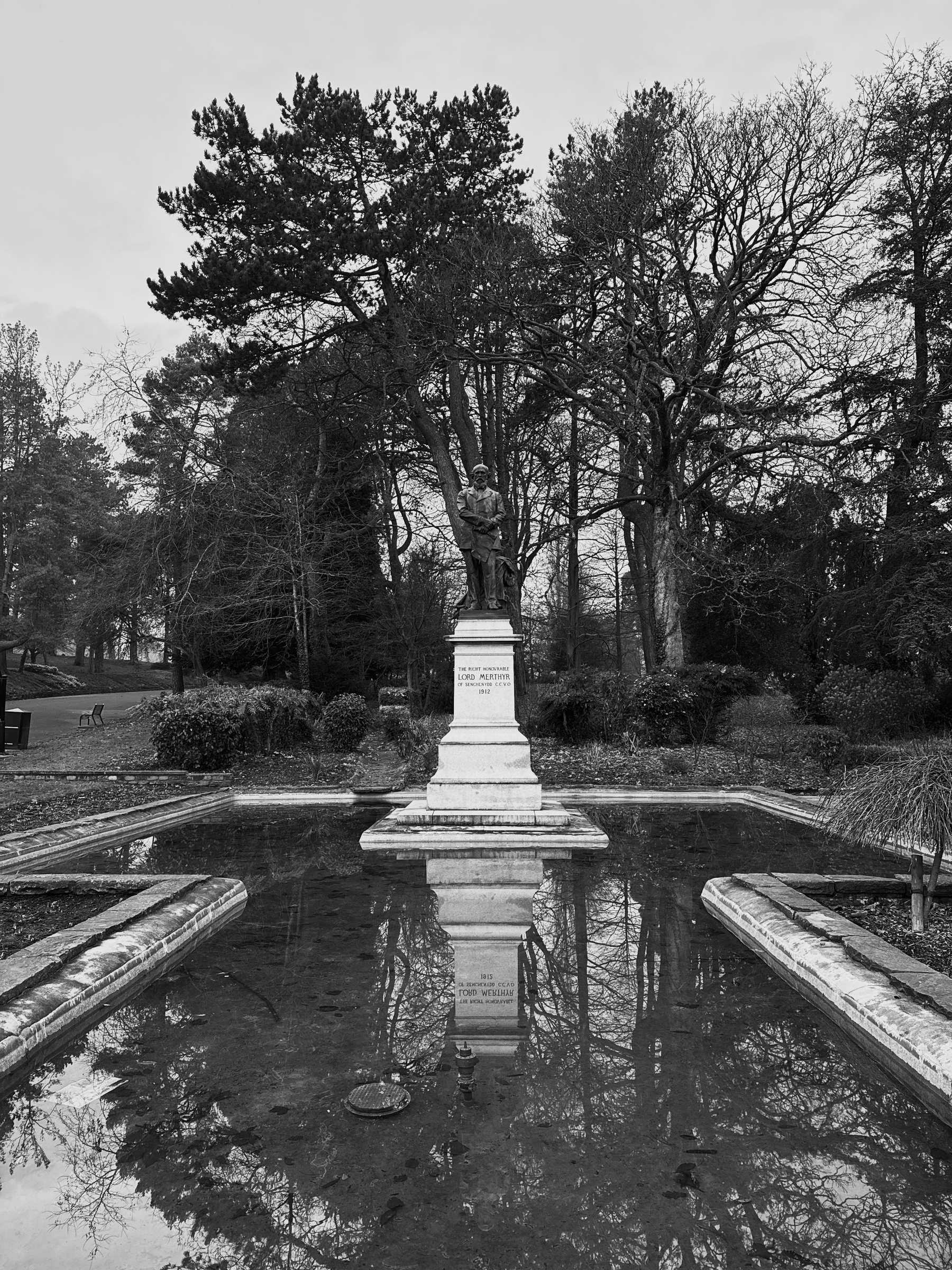 Auto-generated description: A statue stands on a plinth surrounded by a reflective pond, with trees and a pathway in the background.