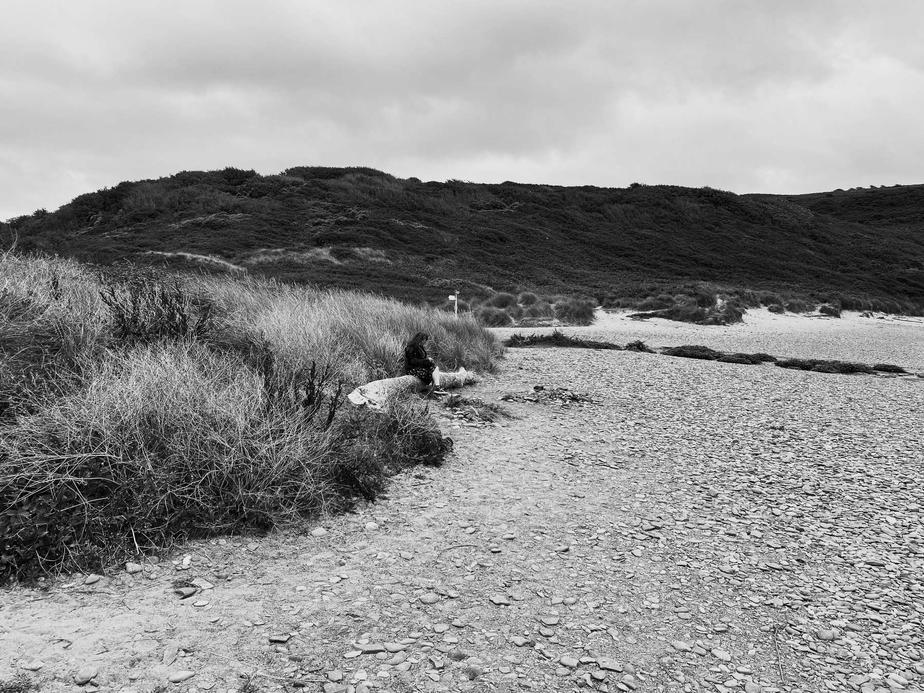 Auto-generated description: A person sits on a rocky path surrounded by grassy dunes and hills under a cloudy sky.